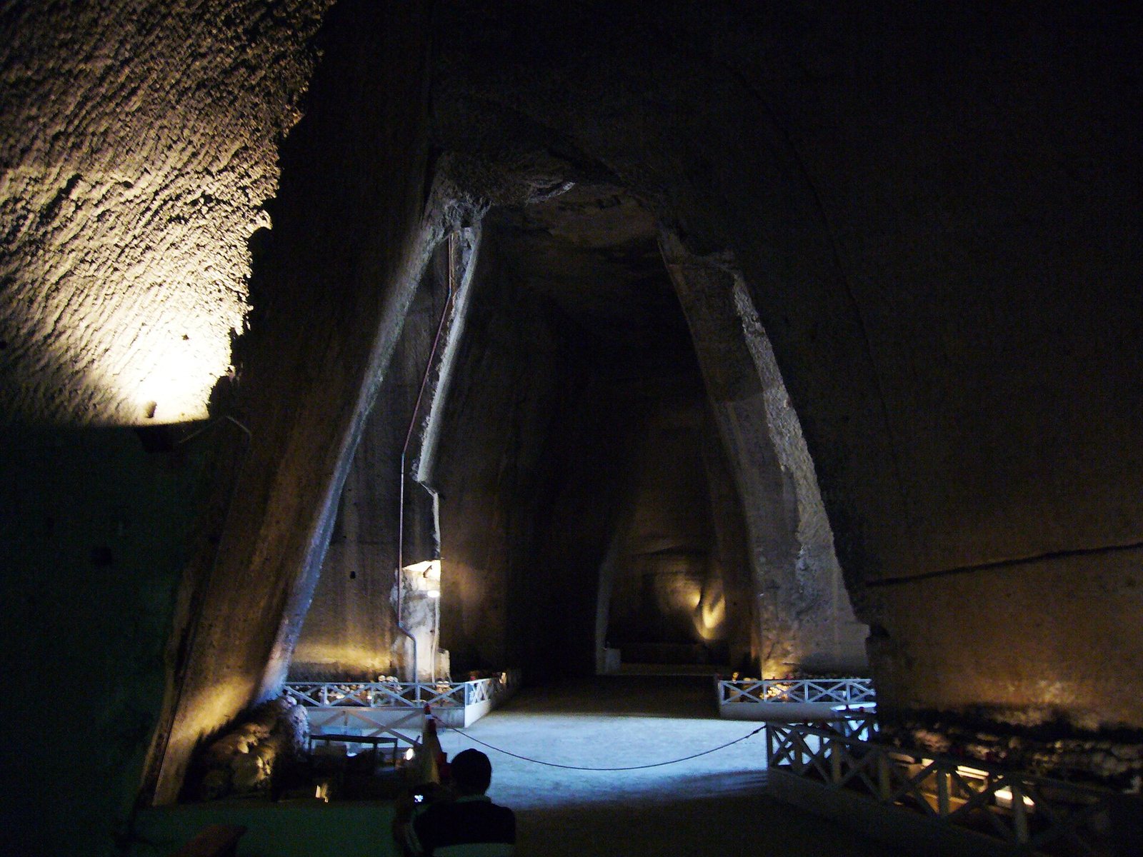 A dimly lit junction of tall quarry galleries in the Fontanelle cemetery, with sloping tuff walls, lamps glowing at the base of the stone and railings marking the visitor route