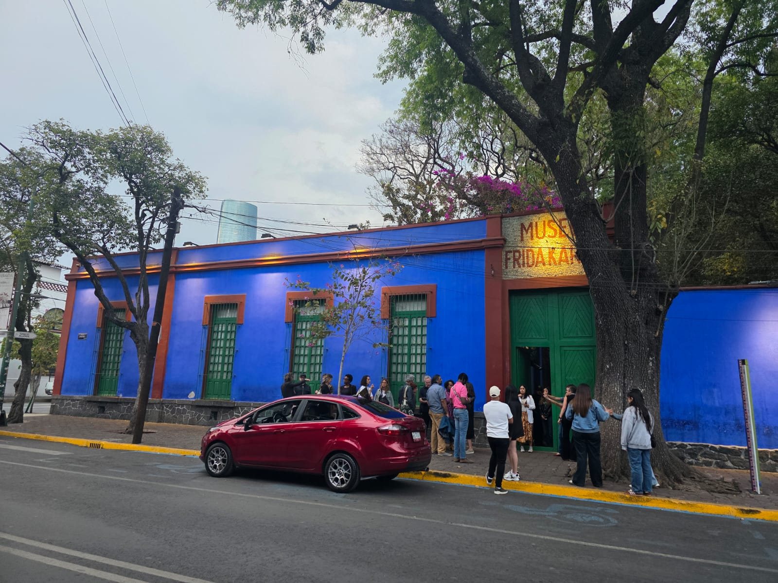 The blue street front of the Museo Frida Kahlo in Coyoacán, with a painted museum sign over the green entrance door, a line of visitors on the sidewalk and a red car parked in the street