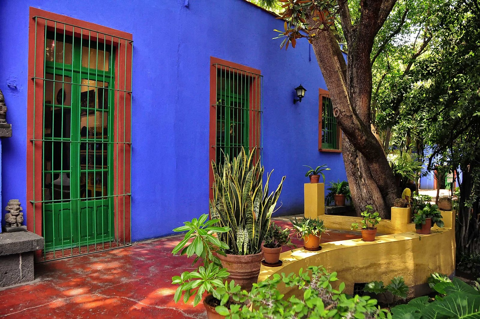 The cobalt-blue wall of the Casa Azul in Coyoacán with a green barred door and window framed in terracotta red, plants in pots and a yellow step in the sunlight