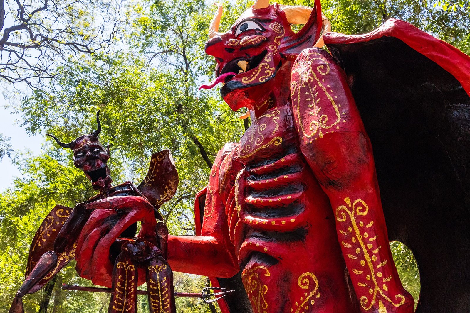 A large red papier-mâché devil figure with horns, wings and gold patterns, holding a smaller horned figure, with green trees behind it