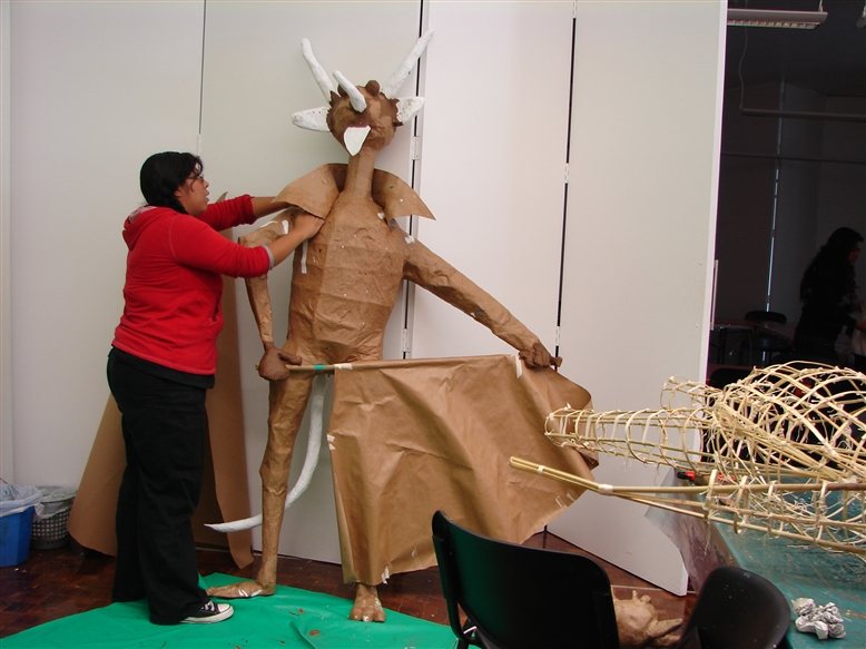 A woman in a red sweater shapes a tall horned figure of brown paper with tusks, beside a wire frame lying on a table in a workshop