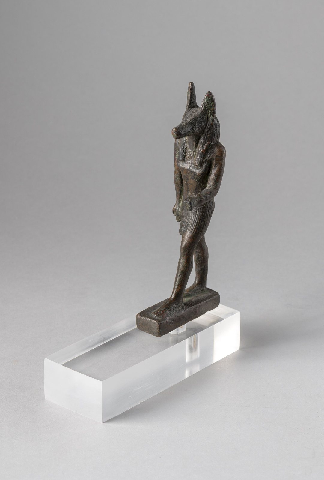 Pequeña estatuilla de bronce oscuro de una figura que avanza, con cuerpo humano y cabeza de chacal de hocico largo y orejas altas, sobre un soporte transparente