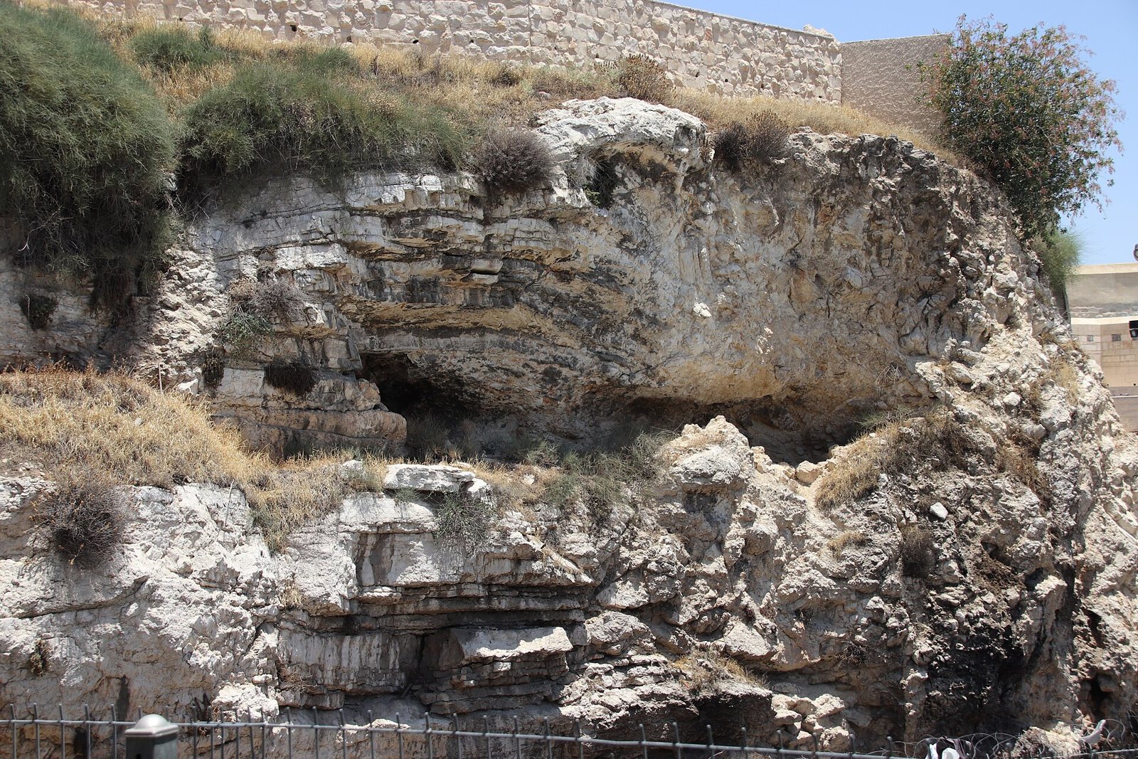Weathered pale limestone cliff with dark hollows and scrub on top, beside a stone wall, known as Skull Hill near the Garden Tomb