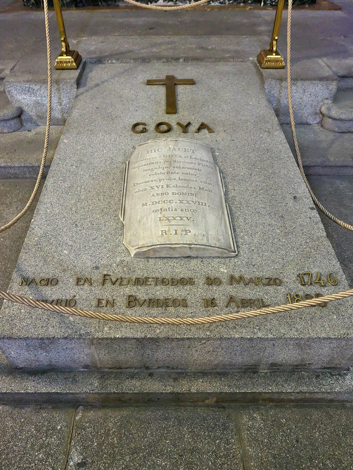 Goya's tomb slab: a granite stone with a cross and the name GOYA above an open stone book, with his birth and death places carved below, behind a gold rope