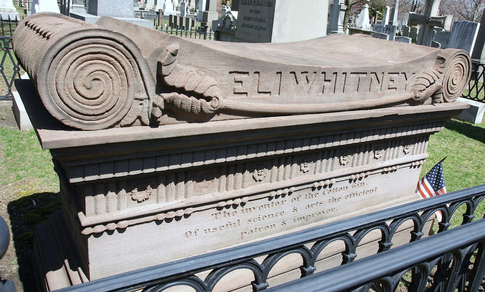 Eli Whitney's stone monument, topped with a carved scroll bearing his name, behind an iron railing