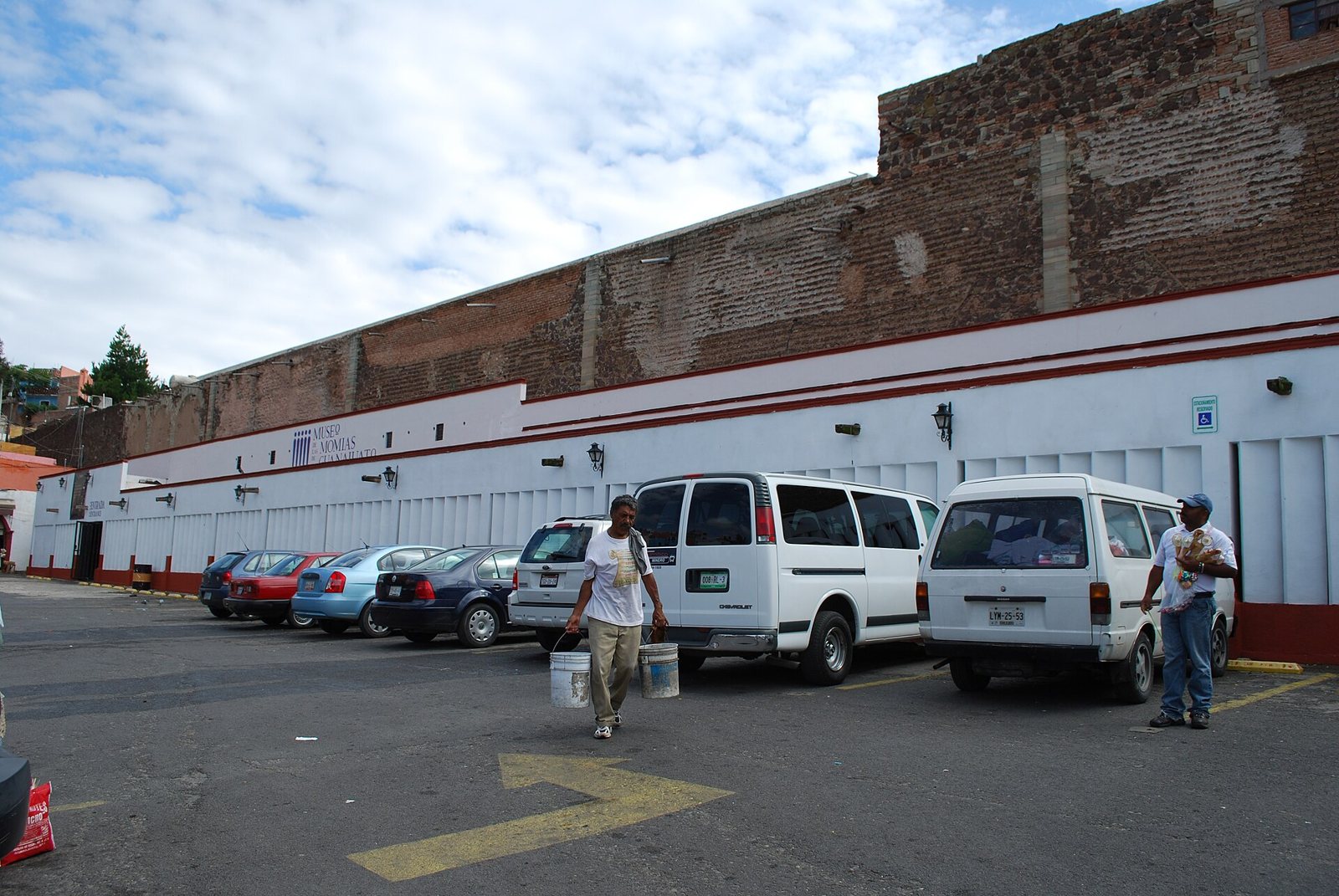 The white and brick-red front wall of the Museo de las Momias with the museum name above the entrance at left, vans and cars parked in front and two men standing in the lot