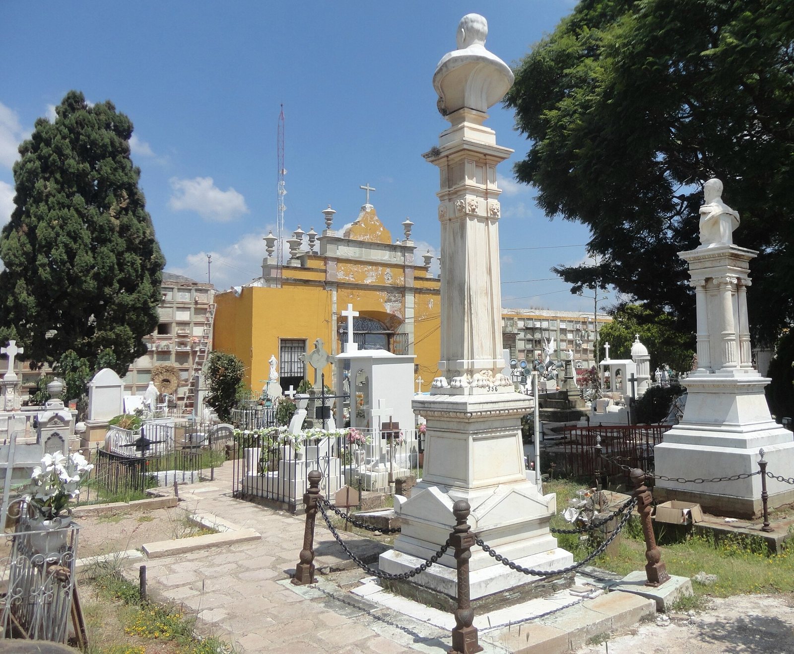 Marble busts on tall pedestals and white tombs in the Santa Paula cemetery, with a yellow chapel and walls of burial niches behind them under a blue sky