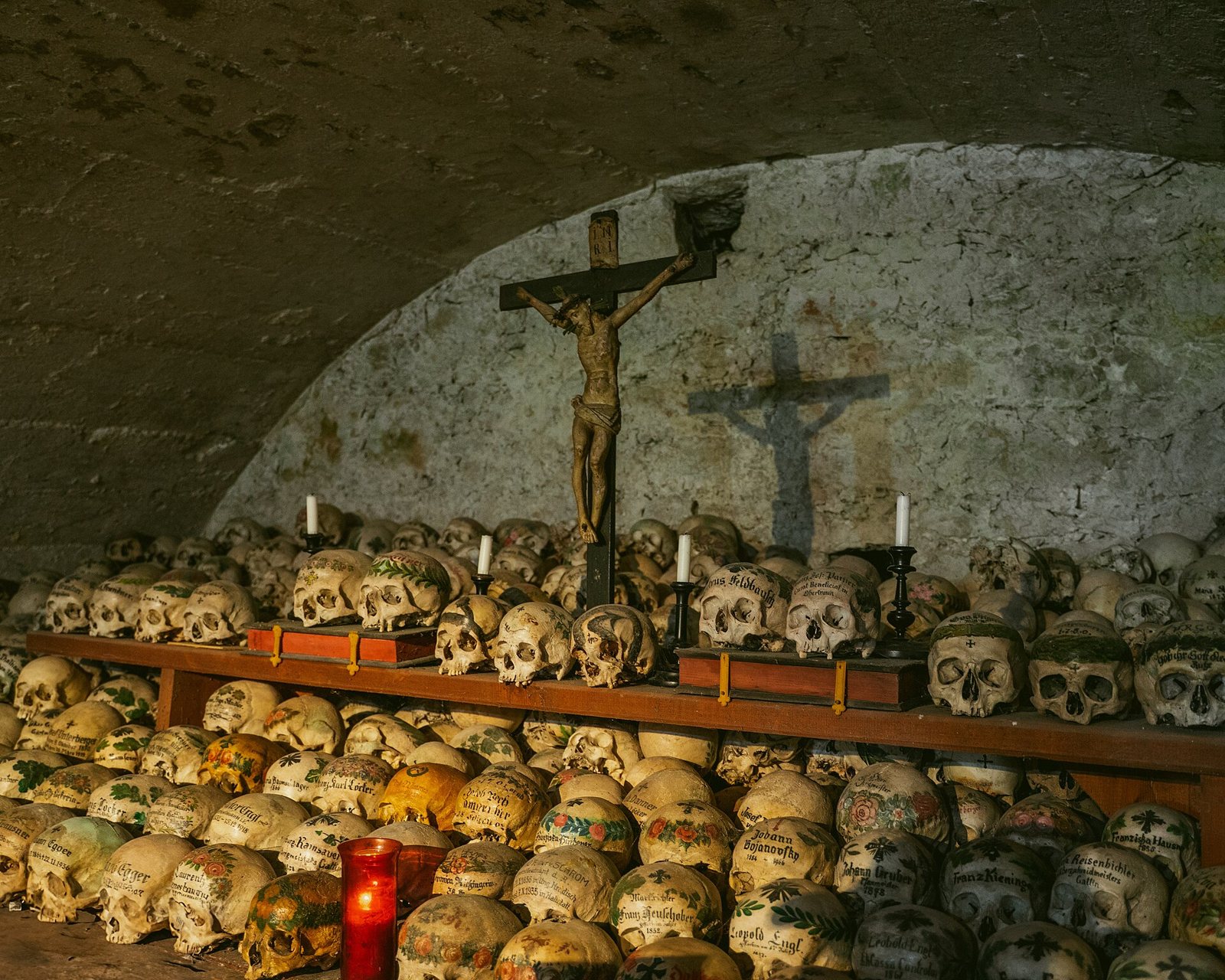 Painted skulls in rows on a wooden shelf beneath a carved crucifix and candles in the Hallstatt Bone House