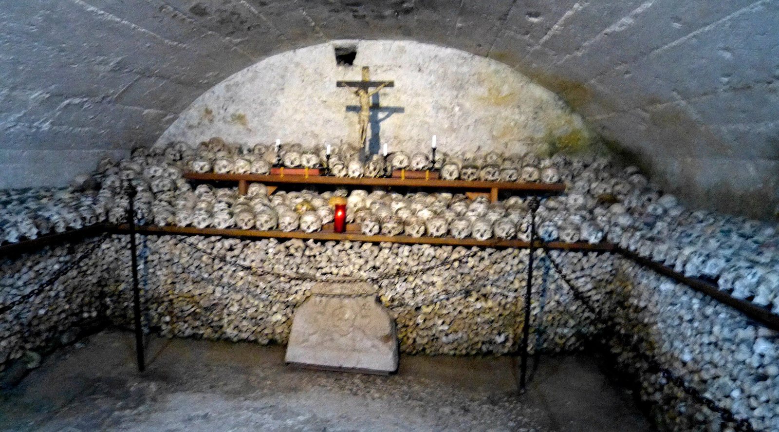The vaulted Bone House in Hallstatt, its walls stacked with skulls and long bones on wooden shelves around a crucifix, with a red votive candle, behind a low chain barrier