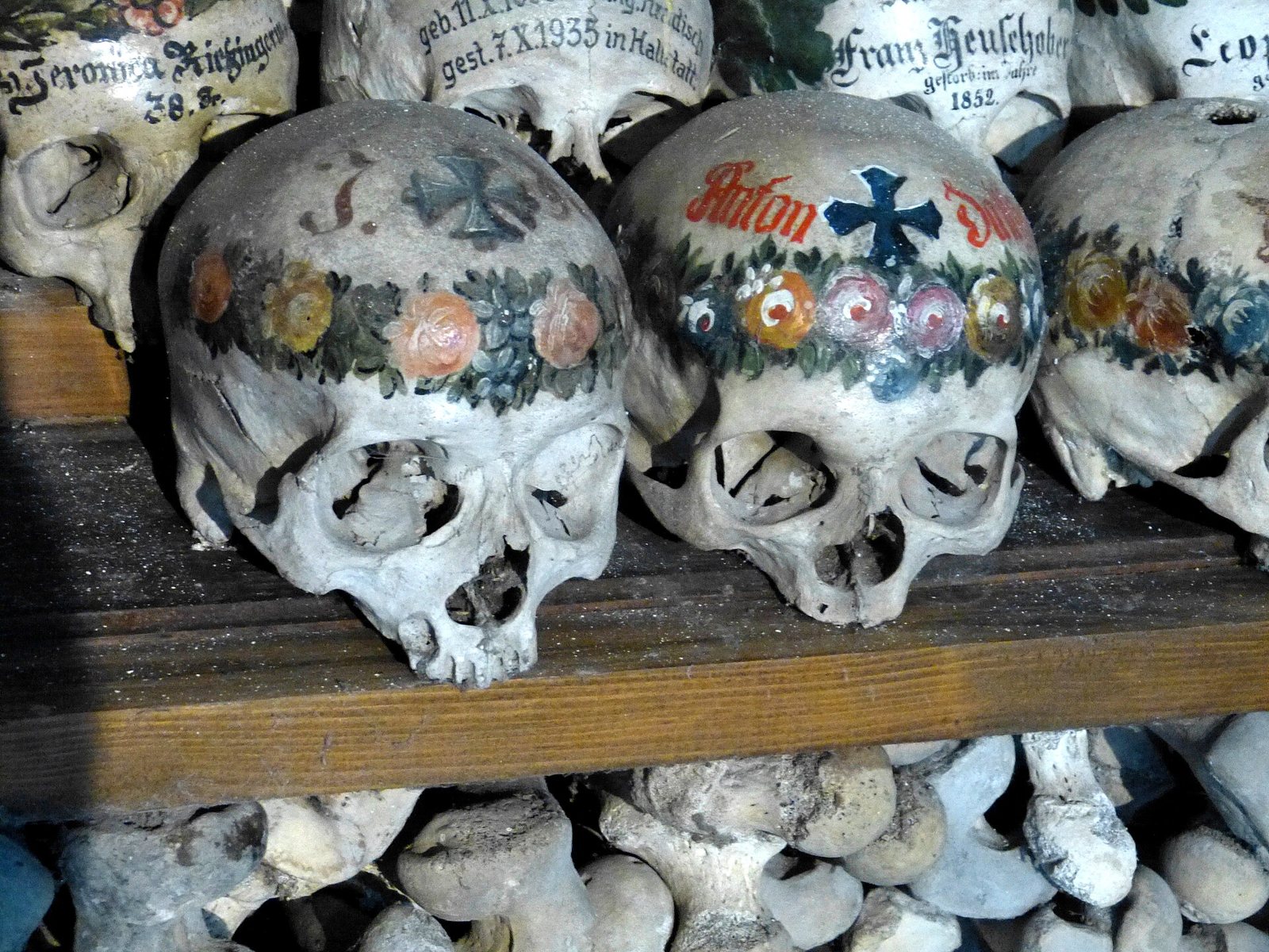 Close view of Hallstatt skulls painted with wreaths of red and pink flowers, dark blue and black crosses, names and death dates, resting on a shelf above stacked bones