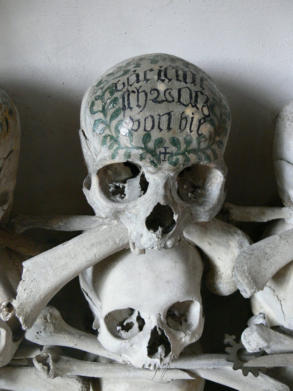 A skull painted with a green leafy wreath, a small cross and lines of black Gothic lettering, resting on crossed long bones above another skull