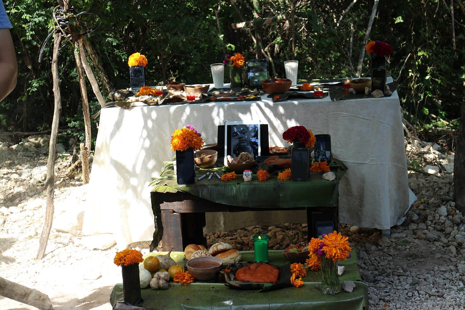 An outdoor Hanal Pixán altar under trees: a table with a white cloth holding cups and dishes, and in front a lower table covered in green leaves with photographs, marigolds, candles, squash and bowls of food