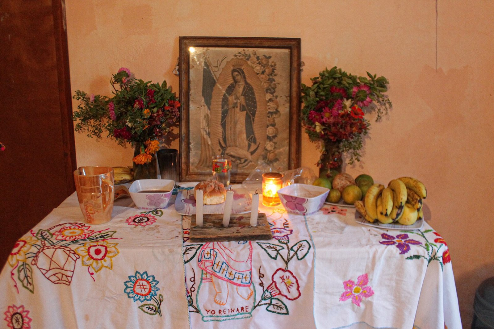 A Hanal Pixán table altar against an orange wall: a framed image of the Virgin of Guadalupe between vases of flowers, with candles, glasses, bowls, bread and bananas on a white cloth embroidered with flowers