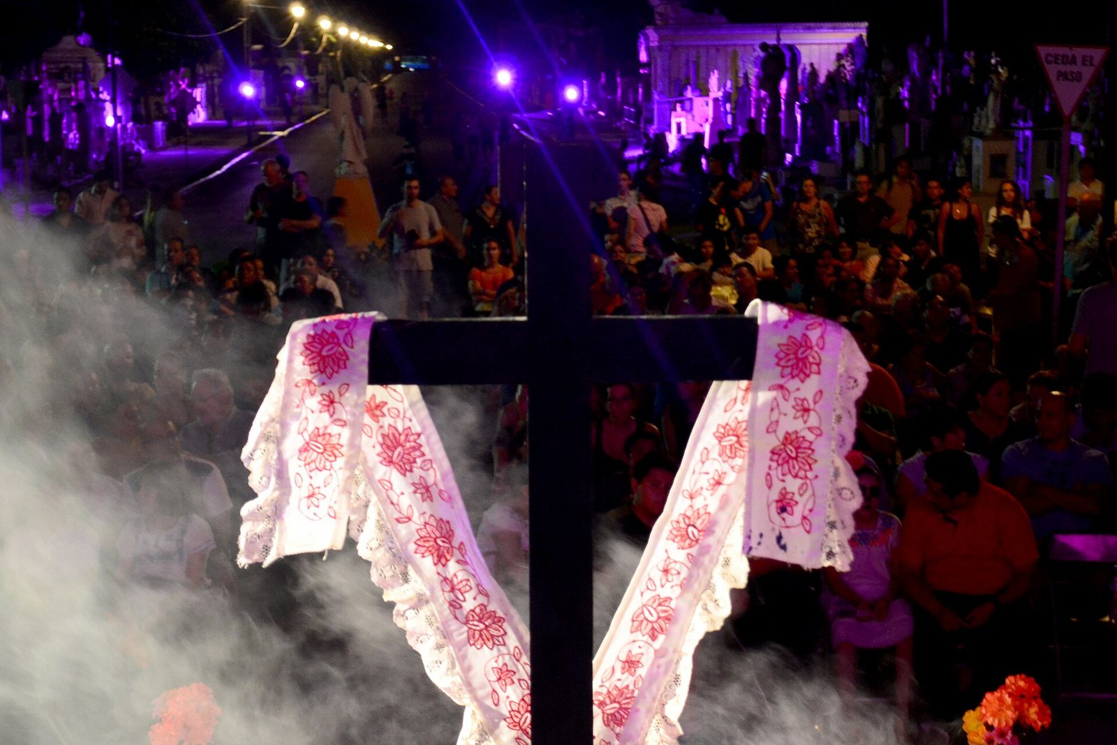 A dark wooden cross draped with an embroidered white cloth, wreathed in copal smoke, in front of a large crowd gathered at night in Mérida's General Cemetery