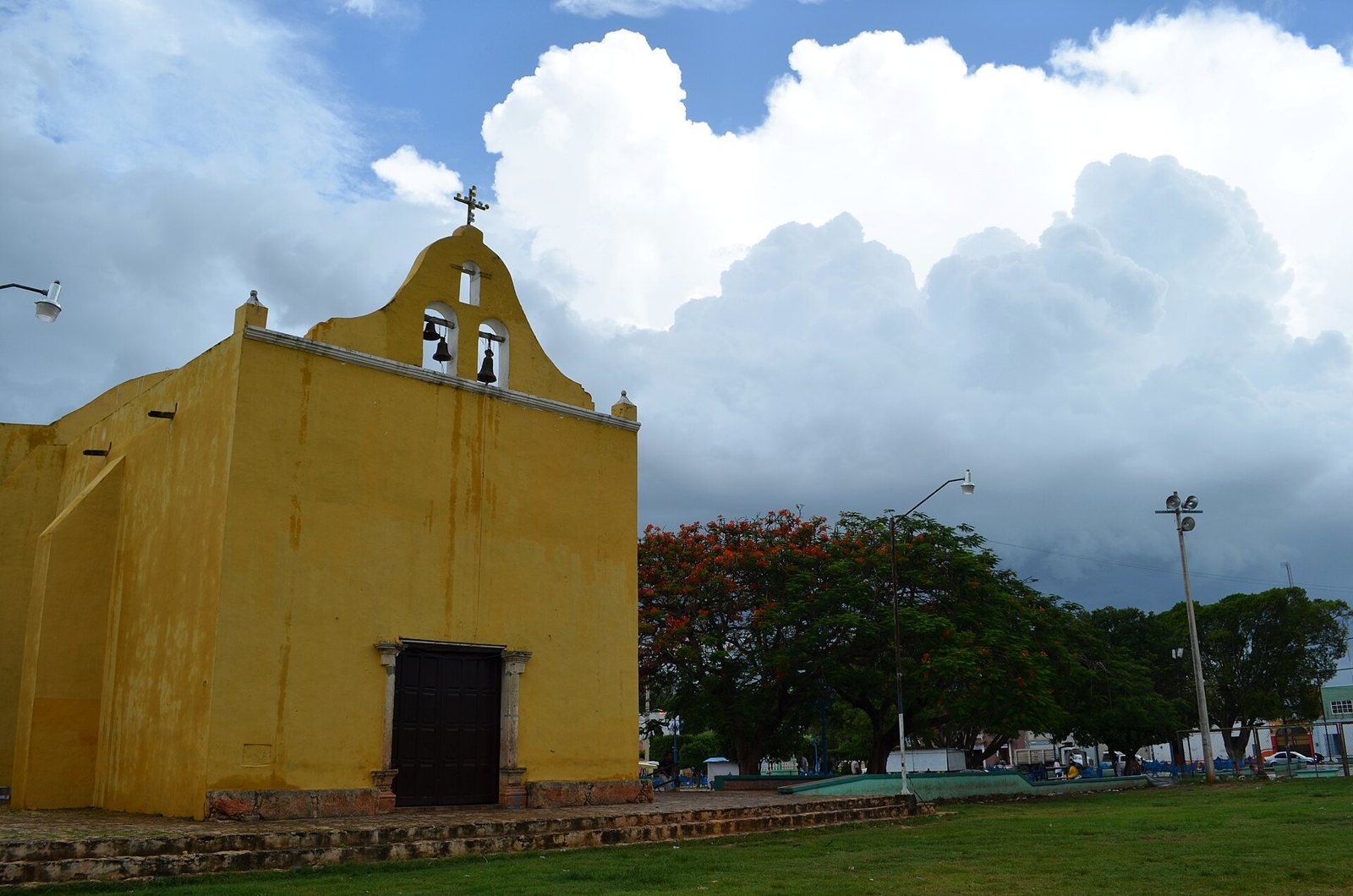 The yellow church of Pomuch, with a stepped bell gable holding two bells, under a sky of tall white clouds beside a lawn and street lamps