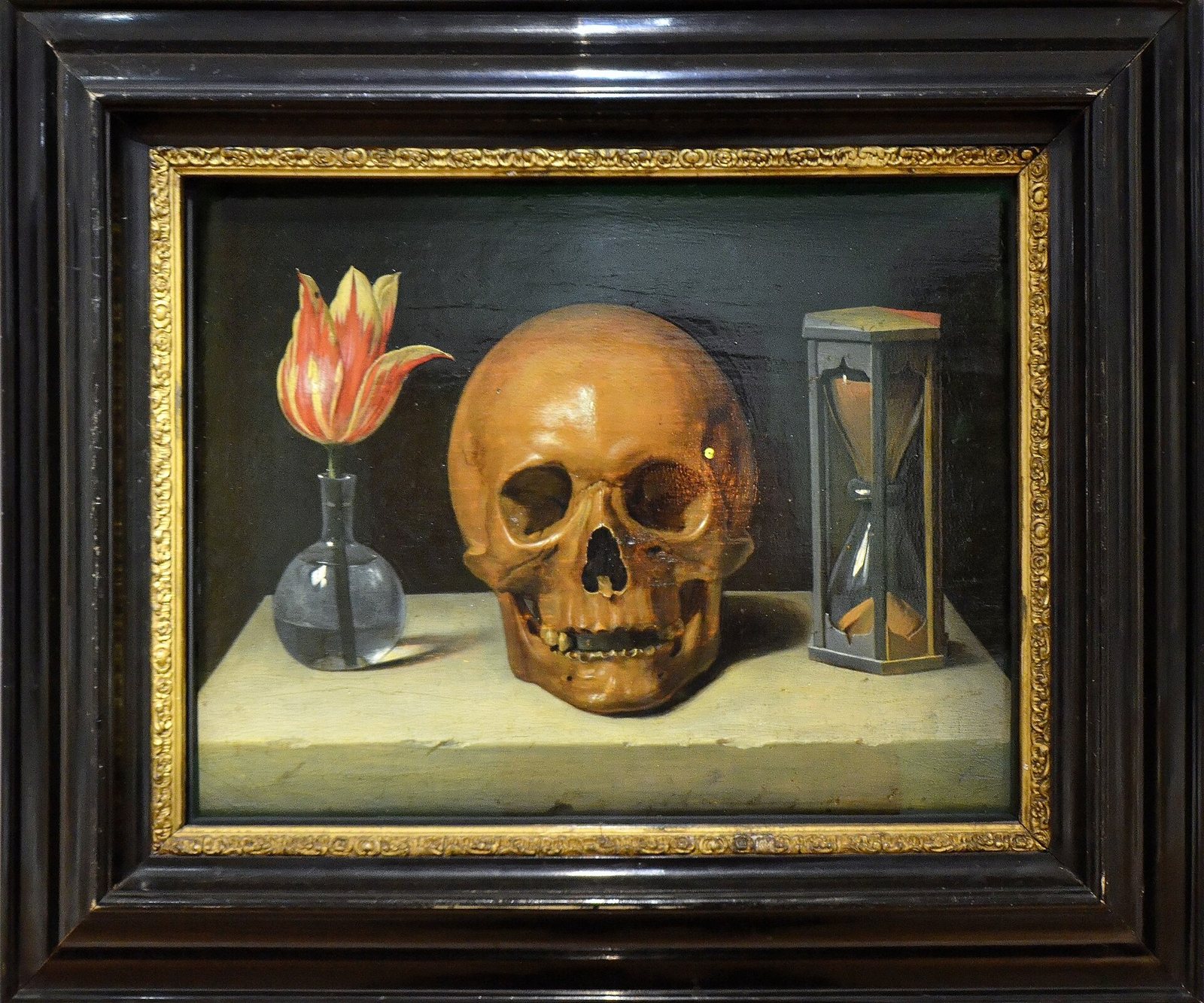A framed painting of a skull on a stone ledge between a red and yellow tulip in a glass vase and an hourglass