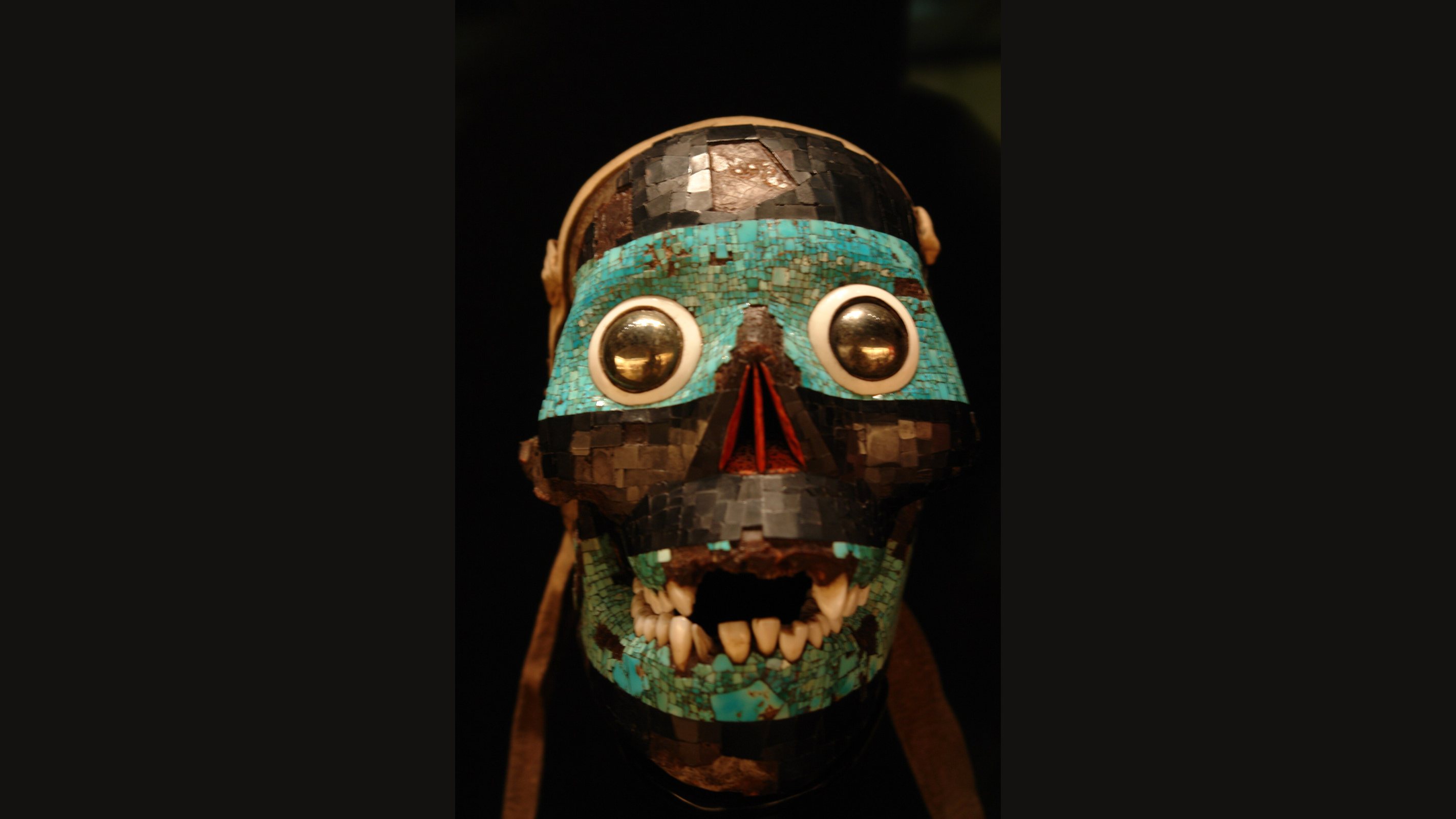 A human skull covered in a mosaic of turquoise bands and dark lignite, with round eyes of white shell and polished pyrite, red strips in the nose opening and real teeth, against a black background