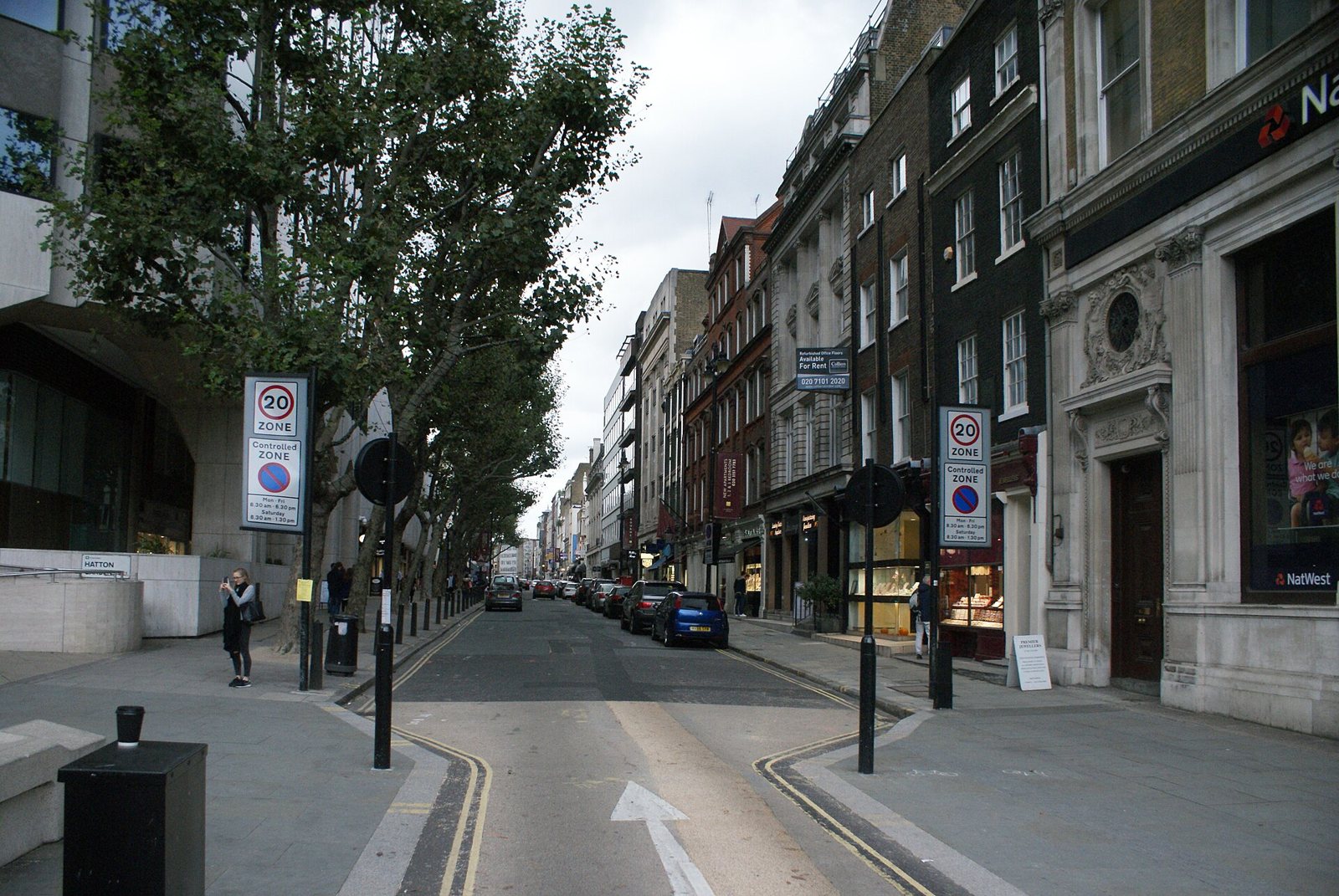 Hatton Garden in London seen from Holborn Circus: a street of stone and brick buildings lined with trees, signs and parked cars