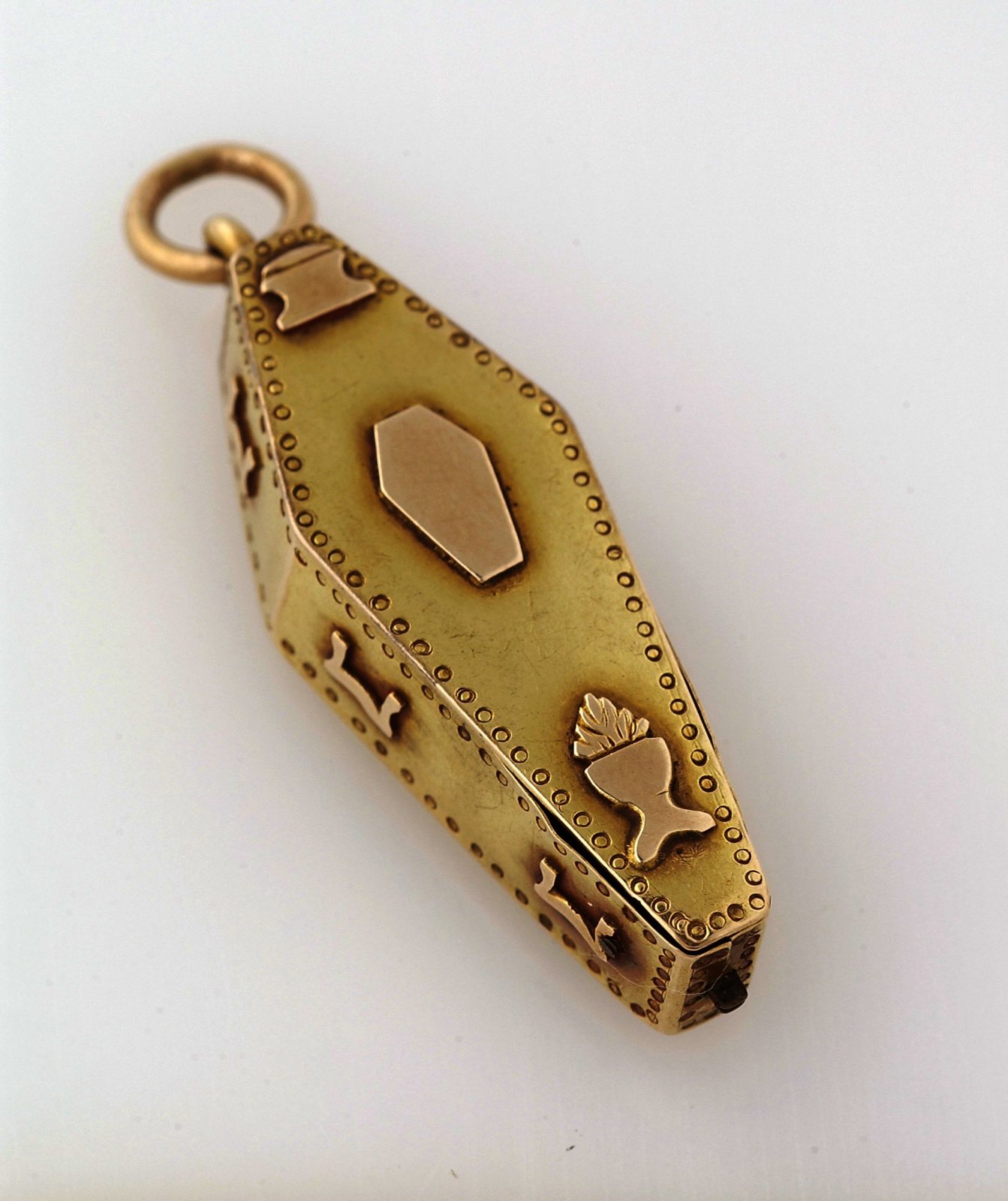A small closed gold pendant shaped like a coffin, with a dotted border and raised rose-gold crossbones, a smaller coffin and a flaming urn on its lid, hanging from a ring