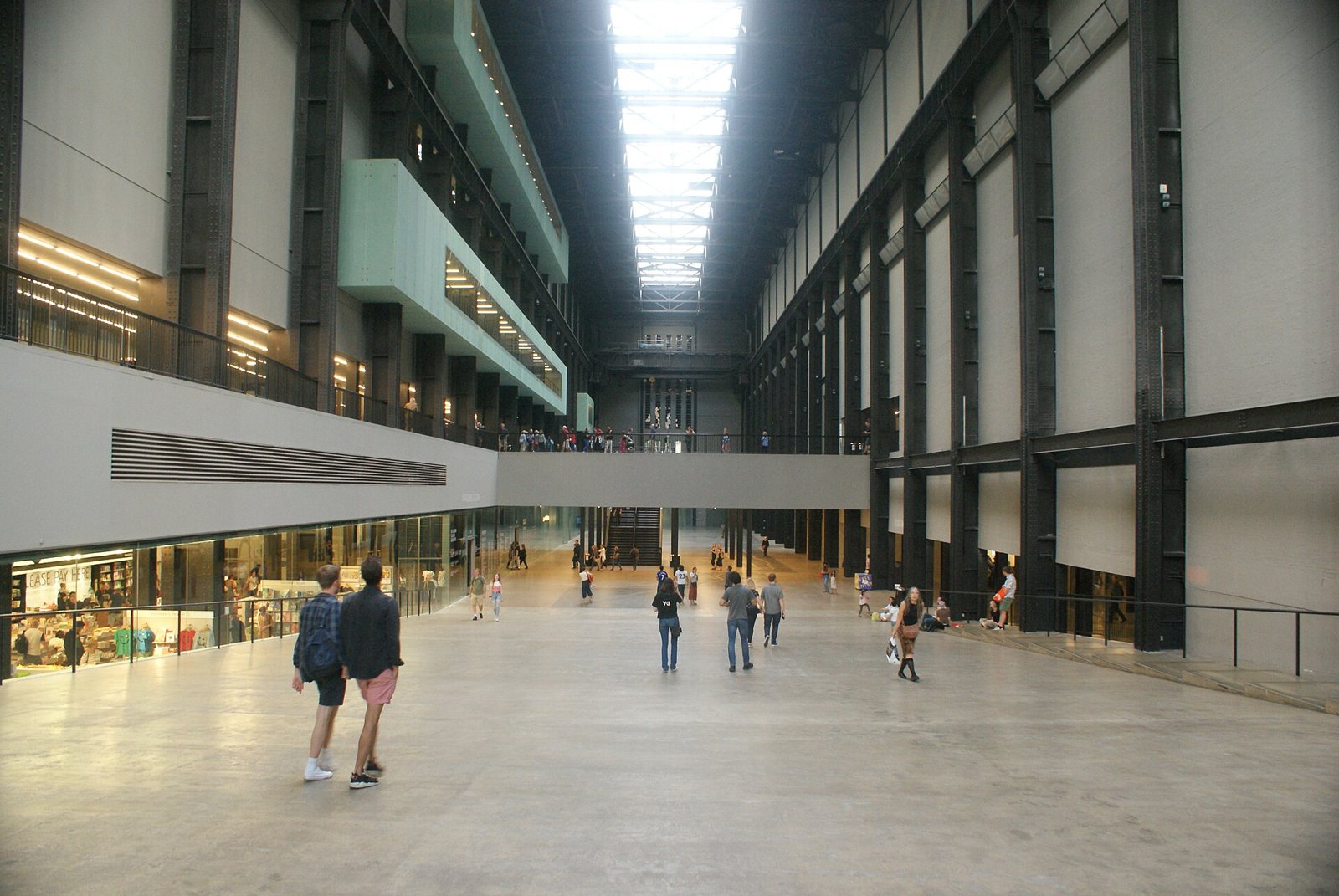 The long, high Turbine Hall at Tate Modern with a skylit roof, steel columns and a few visitors walking across the polished floor