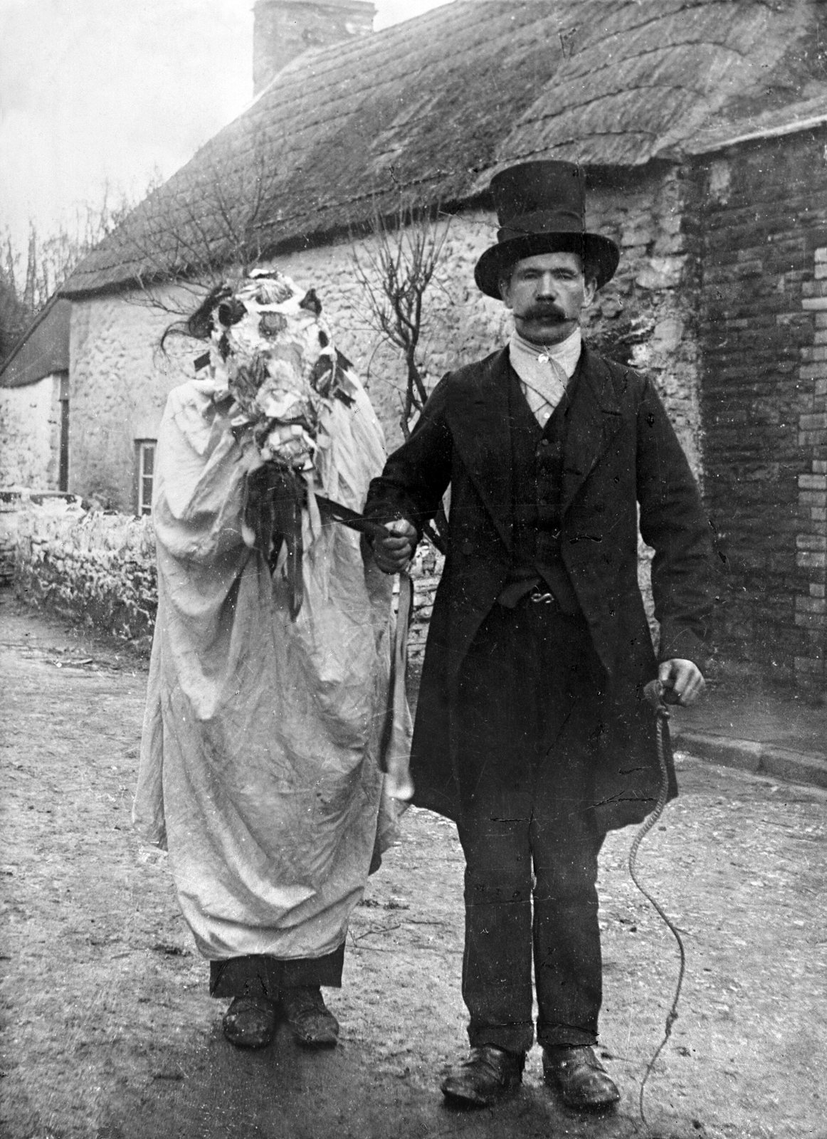Black-and-white photograph of a Mari Lwyd in a village lane: a figure under a white sheet holds a flower-decked skull in front, led by a man in a top hat and dark coat, with a thatched cottage behind