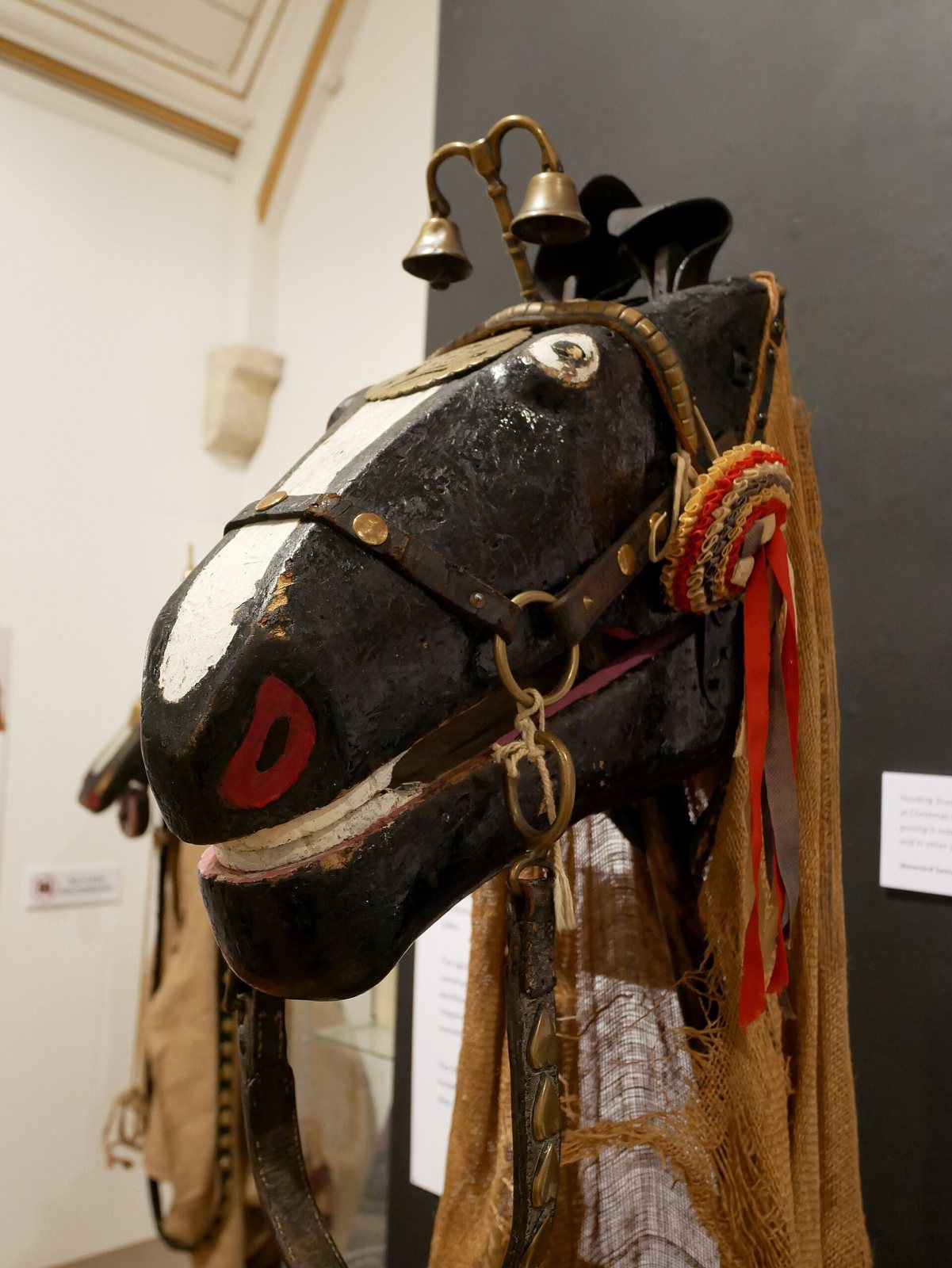 A black wooden Hooden Horse head in a museum, with a white blaze, a red painted mouth, brass bells on the bridle, a red and white rosette and sacking hanging behind