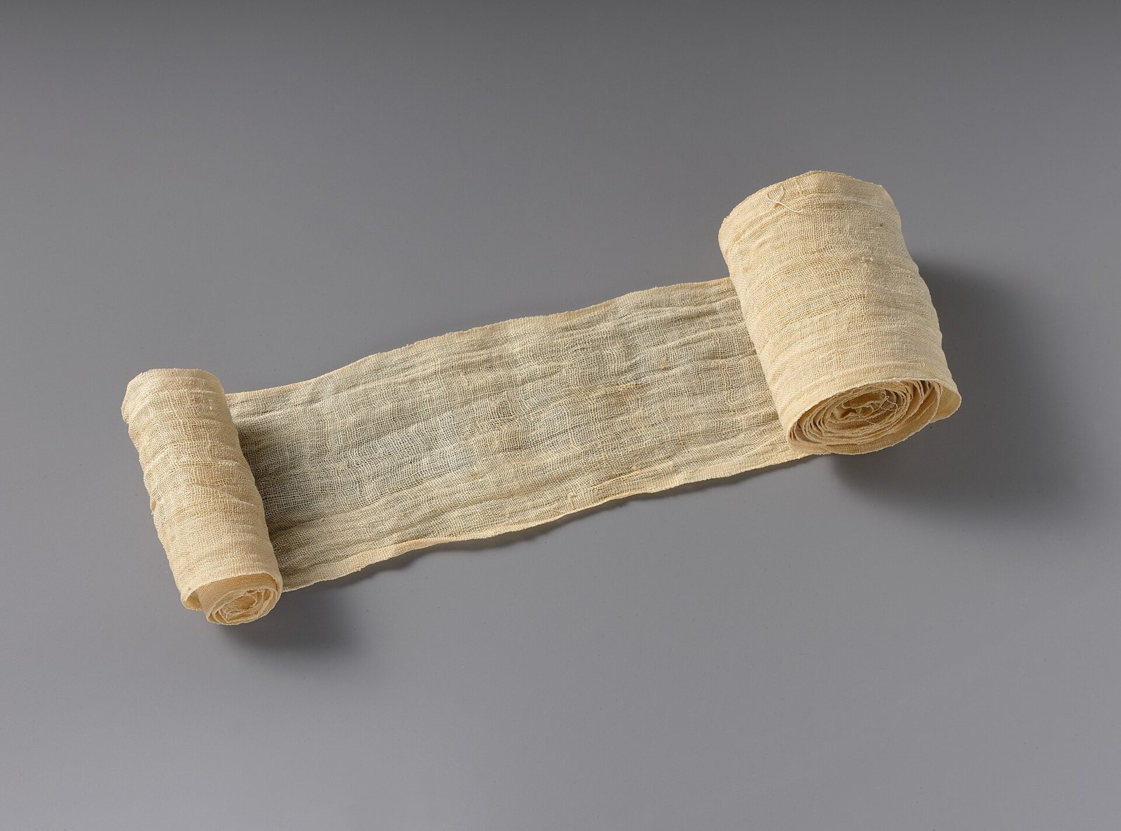 A roll of fine, loosely woven cream linen bandage partly unrolled across a grey surface, with a tightly wound roll at each end