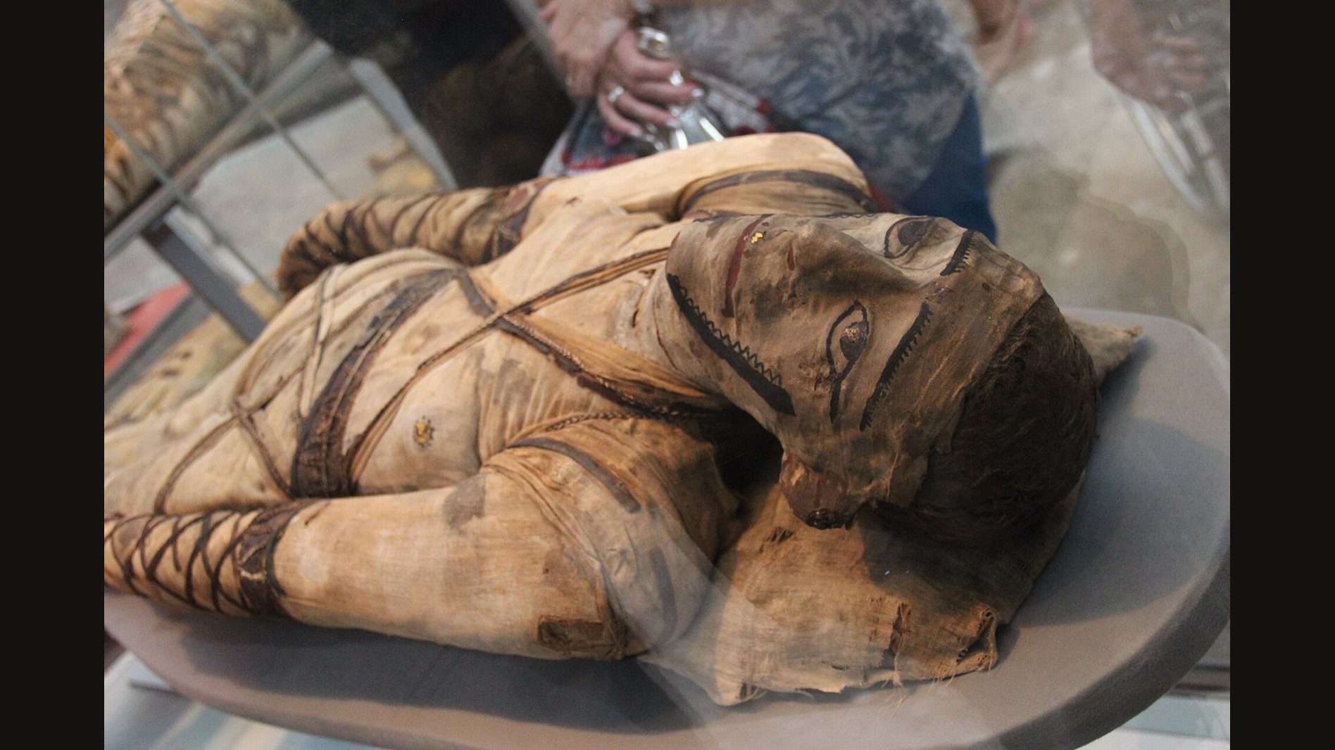 A wrapped mummy lying in a museum case, seen from above and at an angle: linen bandages crossed over the chest, a painted face cover with large outlined eyes, dark hair and bandaged arms