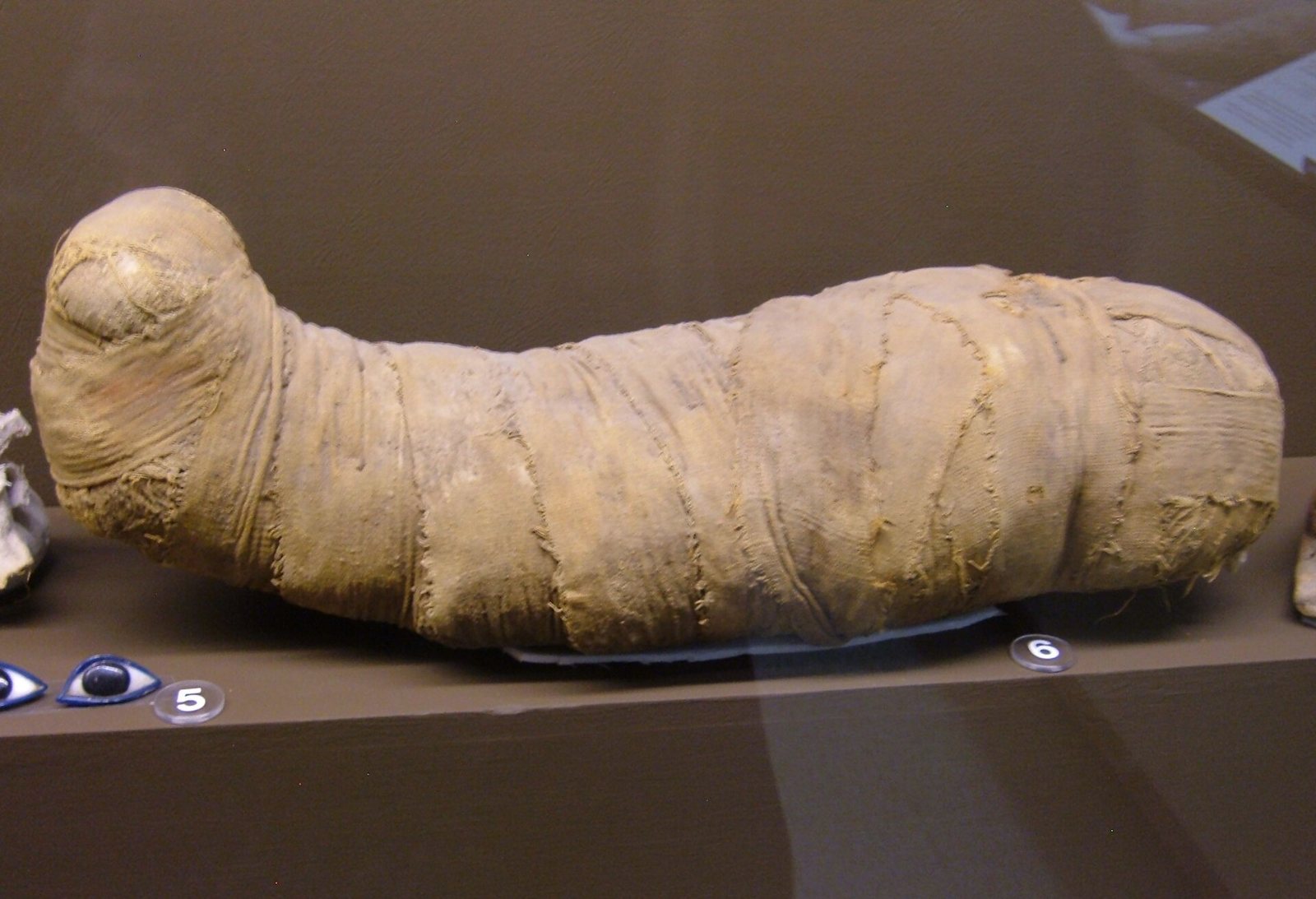 A long bundle of natron wrapped in linen bandages and shaped like a small mummy, lying on a dark museum shelf beside small blue glass eye inlays