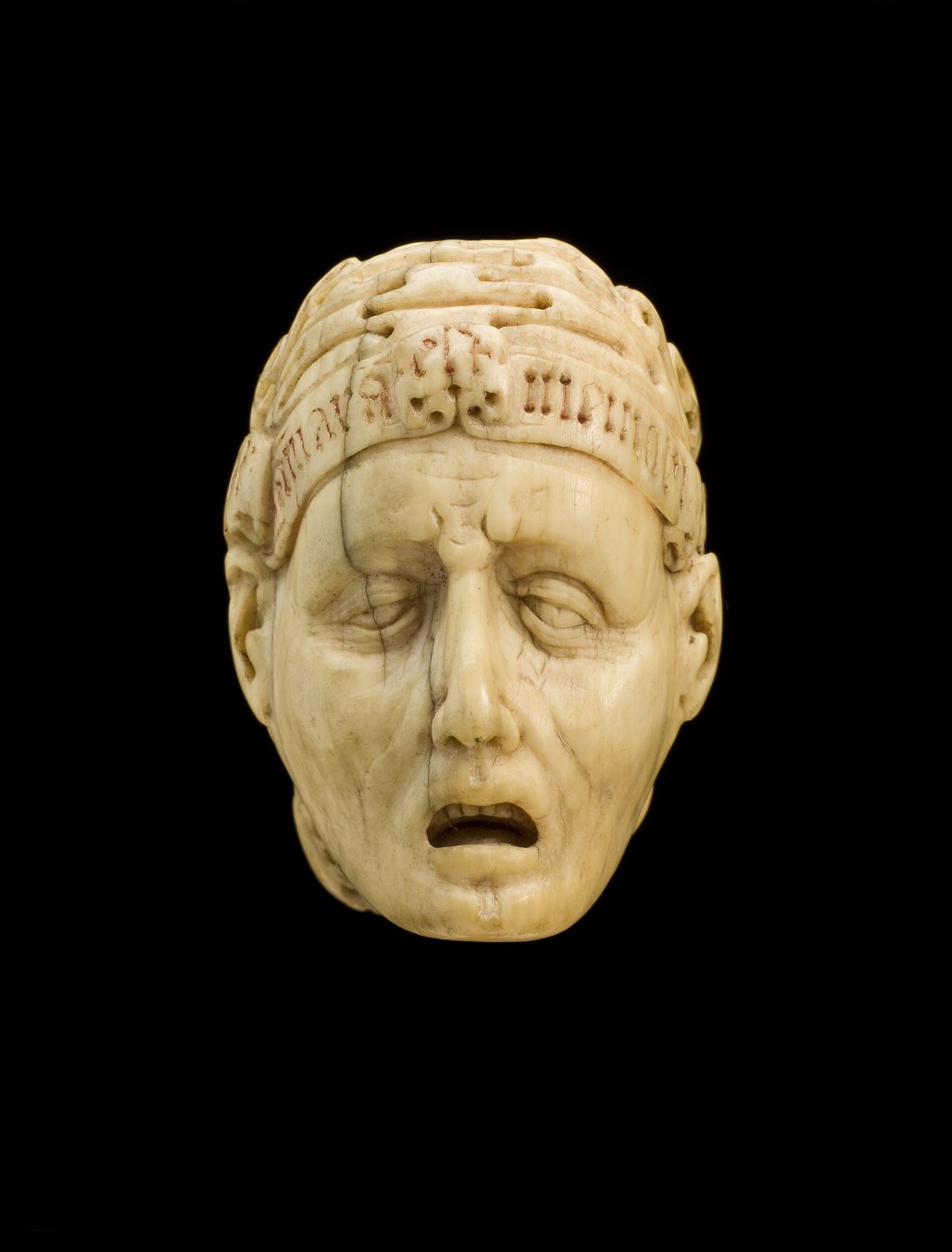 A carved ivory head on a black background, a man's face with closed eyes and an open mouth under an inscribed band; the model is half face and half skull