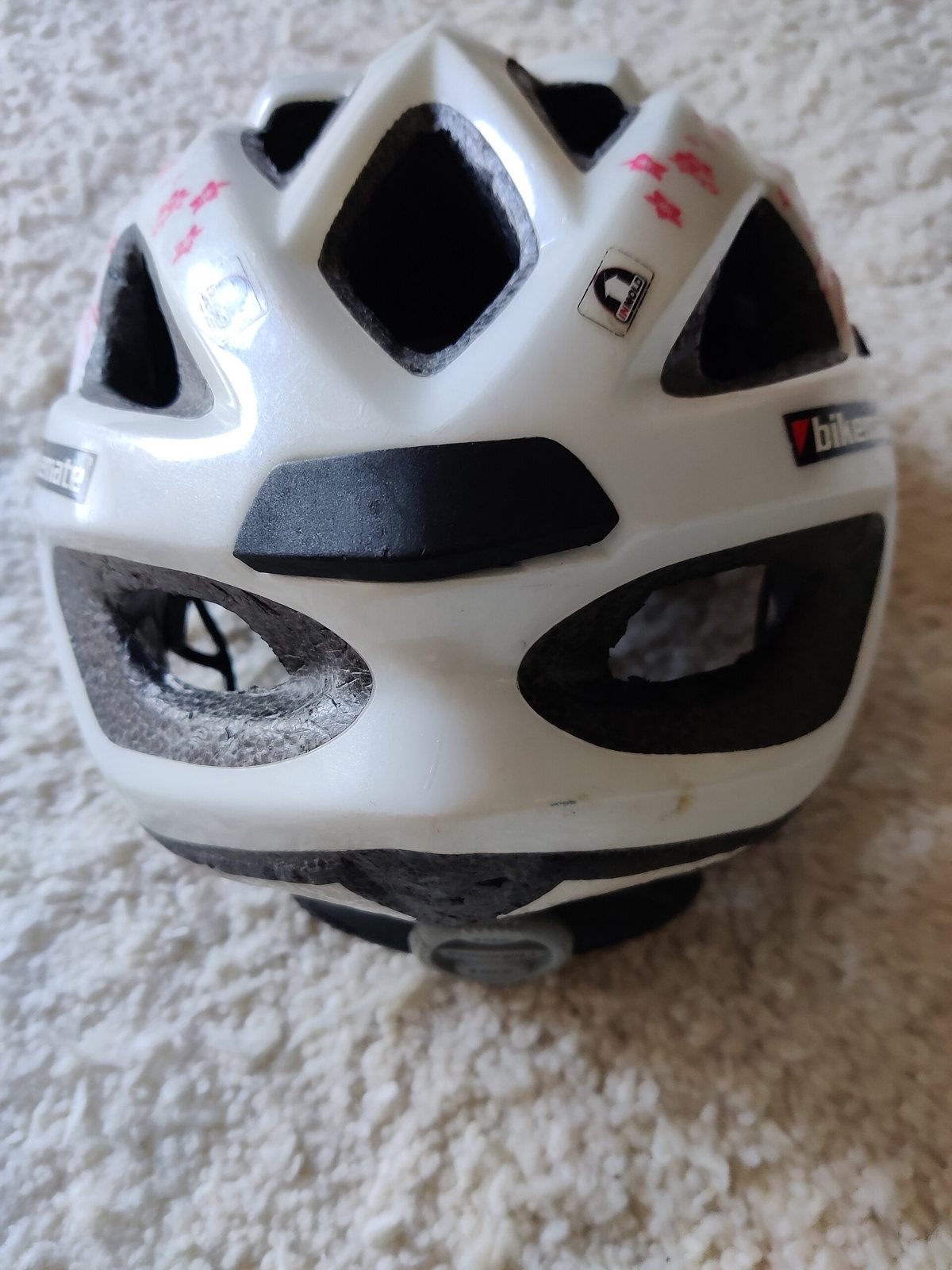 A white bicycle helmet seen from behind on a cream carpet, with vents, black padding and a strap, and the brand name on the shell