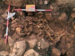 Excavation photograph of the embracing skeletons of Alepotrypa, two people lying together among stones in a trench marked with a scale and a label