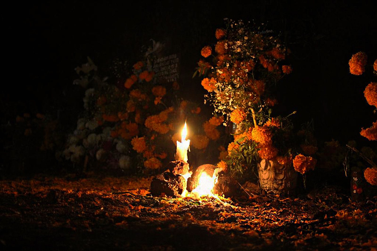 A lit candle on a grave in the dark, with orange marigold flowers piled behind it and scattered petals on the ground
