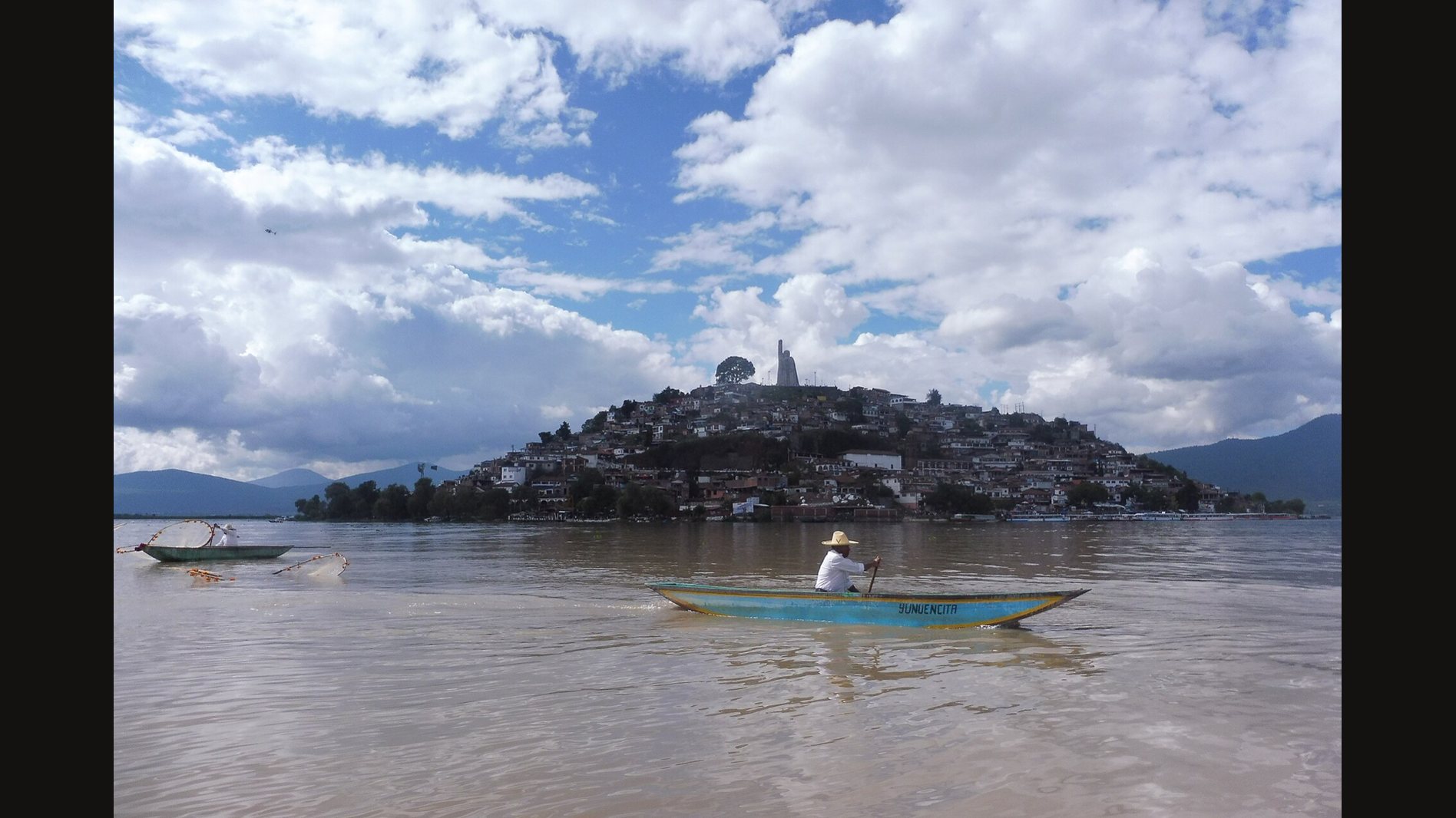 The island of Janitzio on Lake Pátzcuaro under a cloudy sky, its hillside crowded with houses and a tall monument on top, with a fisherman in a small blue boat in the foreground