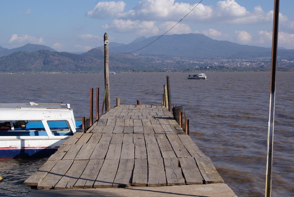 A weathered wooden pier running out into Lake Pátzcuaro, with a white and blue passenger boat tied alongside and mountains across the water