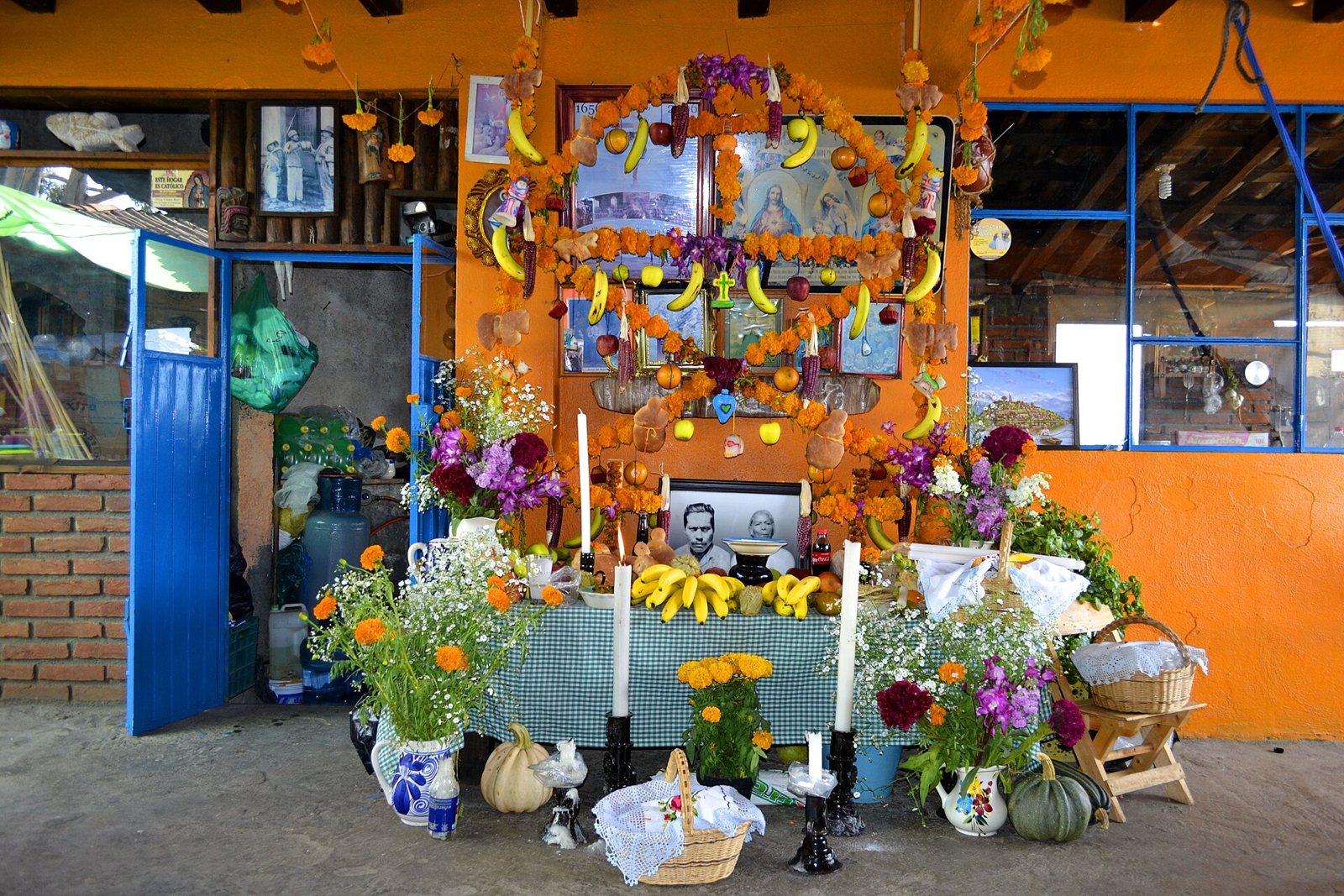 A home ofrenda against an orange wall: an arch of marigolds and bananas around framed photographs, with white candles, bright flowers, squashes and baskets on a table below