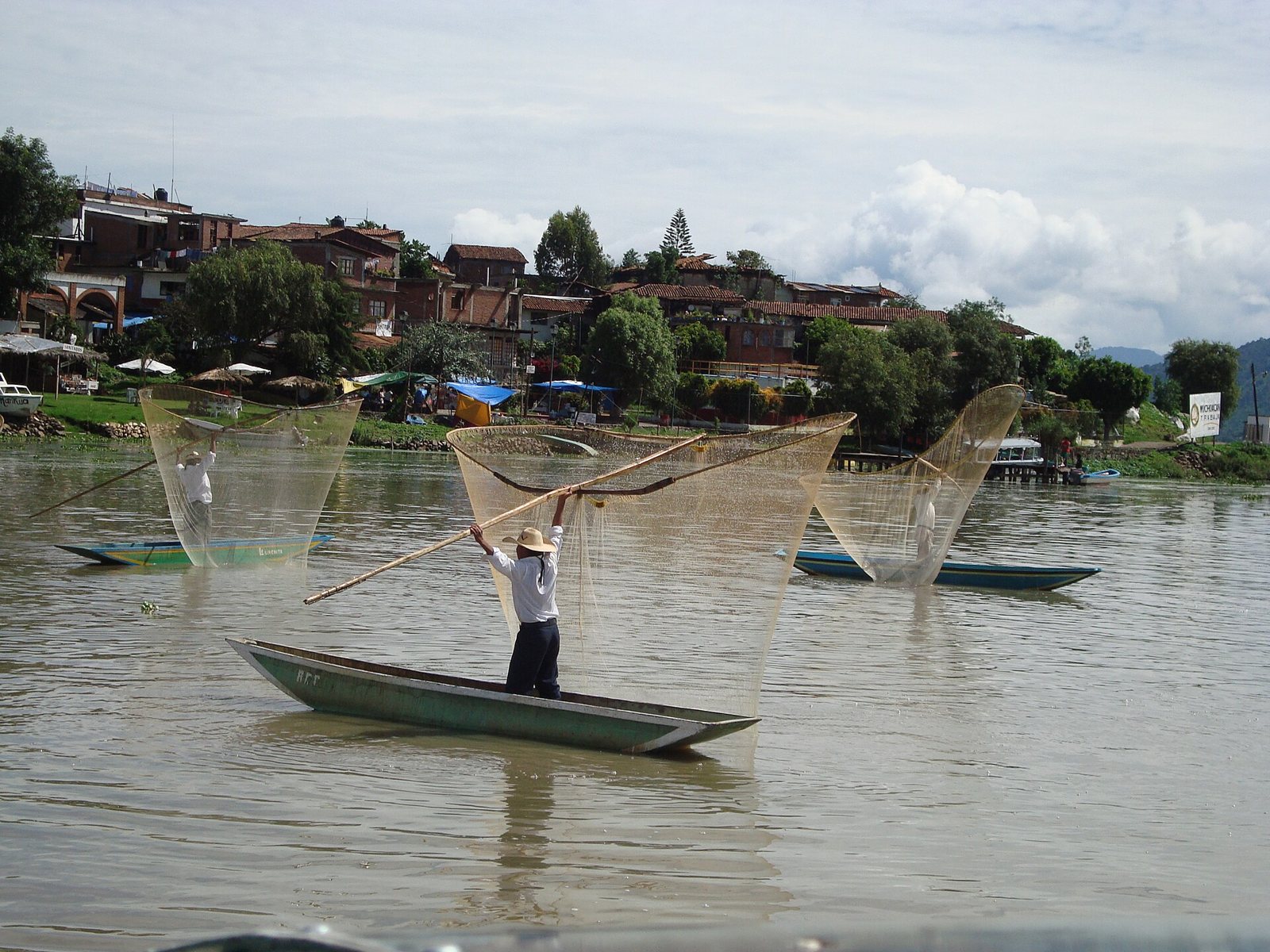 A fisherman standing in a narrow boat on Lake Pátzcuaro, raising a long pole with a wide butterfly-shaped net, with two more nets spread on the water and a lakeside town behind