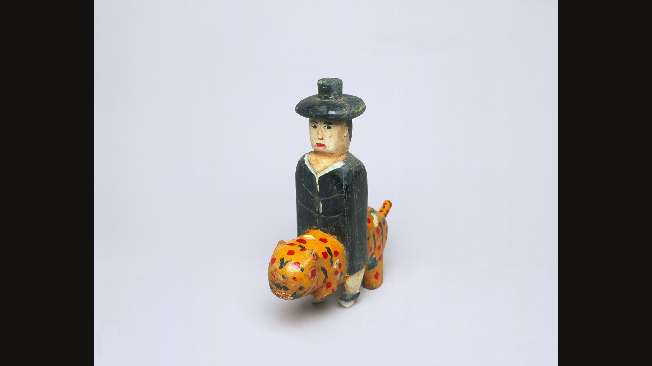 Una pequeña figura de madera pintada de un hombre con abrigo negro y sombrero negro que monta un animal amarillo con manchas rojas, sobre un fondo liso y claro