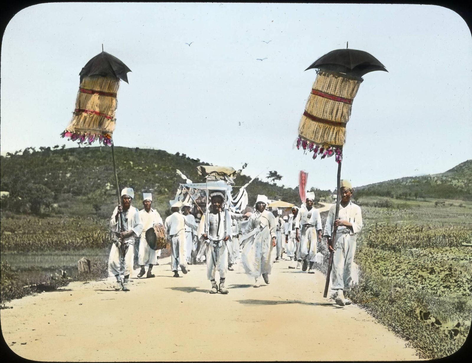 Fotografía coloreada a mano de un cortejo fúnebre en un camino de tierra: hombres de blanco llevan altos estandartes en forma de sombrilla y un tambor, con las andas al fondo y colinas verdes detrás