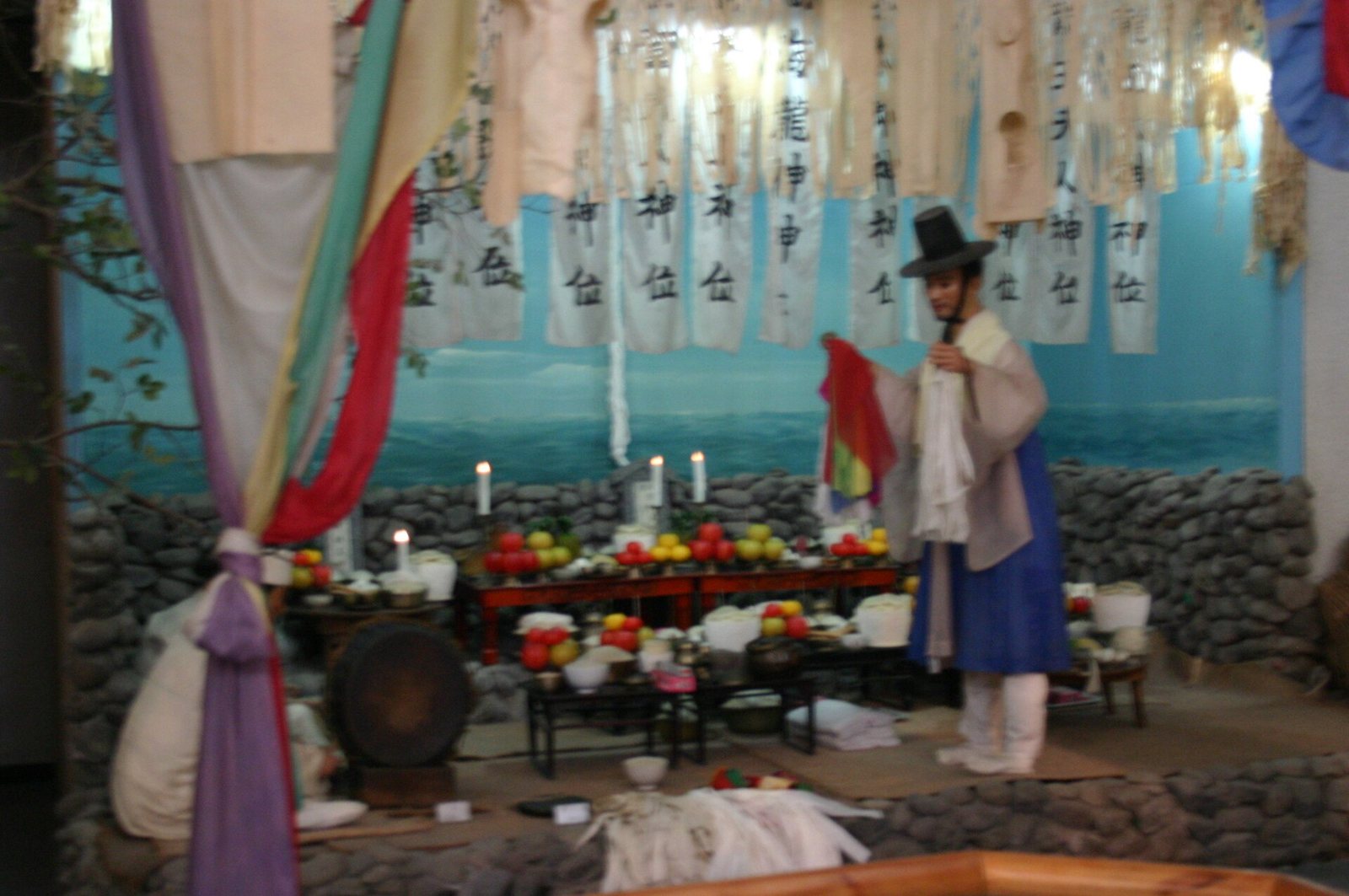 Una exposición de museo de un rito de Jeju: un hombre con sombrero negro y túnica azul está de pie ante un altar con frutas y velas, bajo banderines de papel colgantes con caracteres coreanos