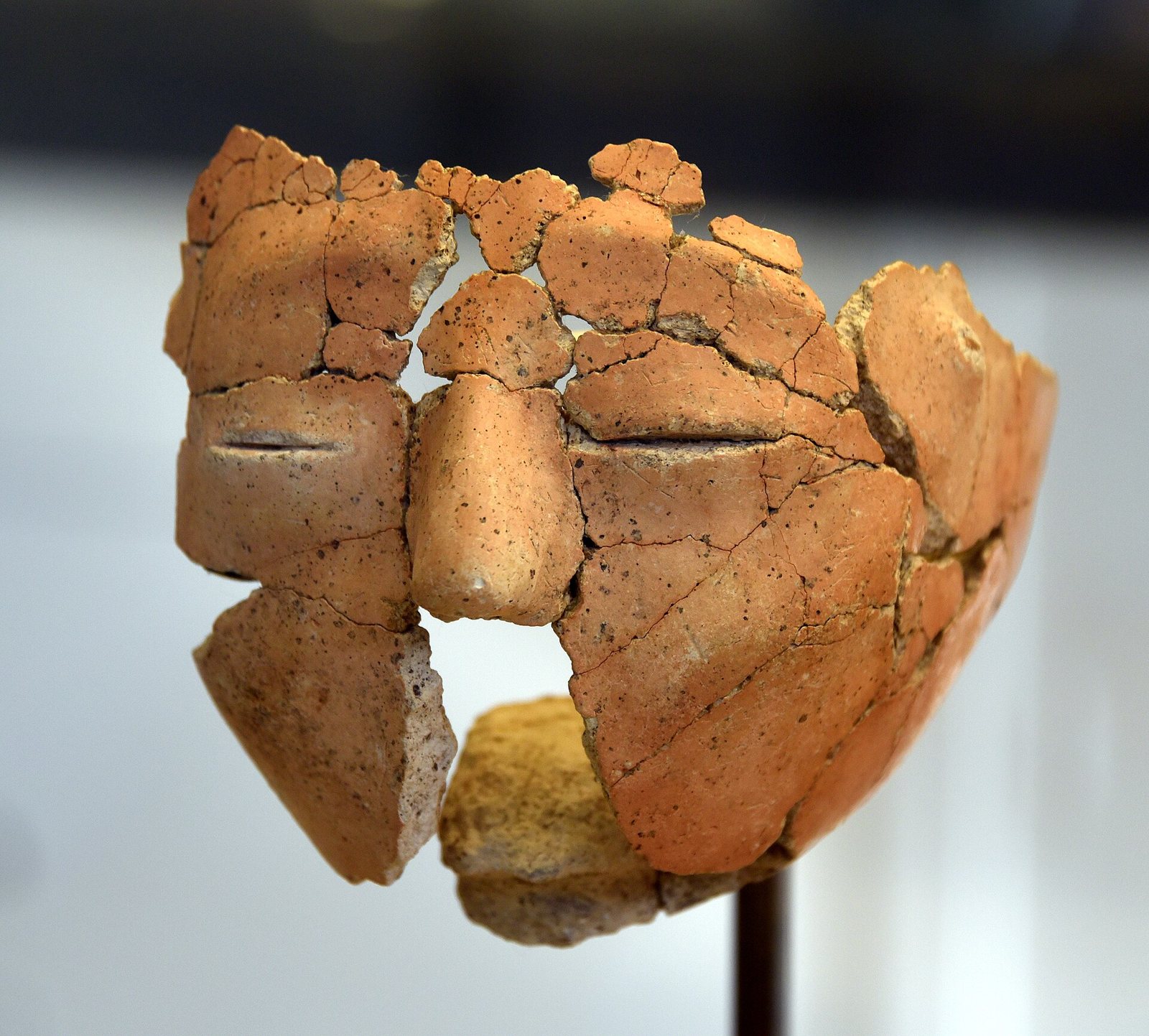 A reassembled plaster face from 'Ain Ghazal on a museum mount, its orange-tan fragments forming closed eyes drawn as slits, a straight nose and cheeks