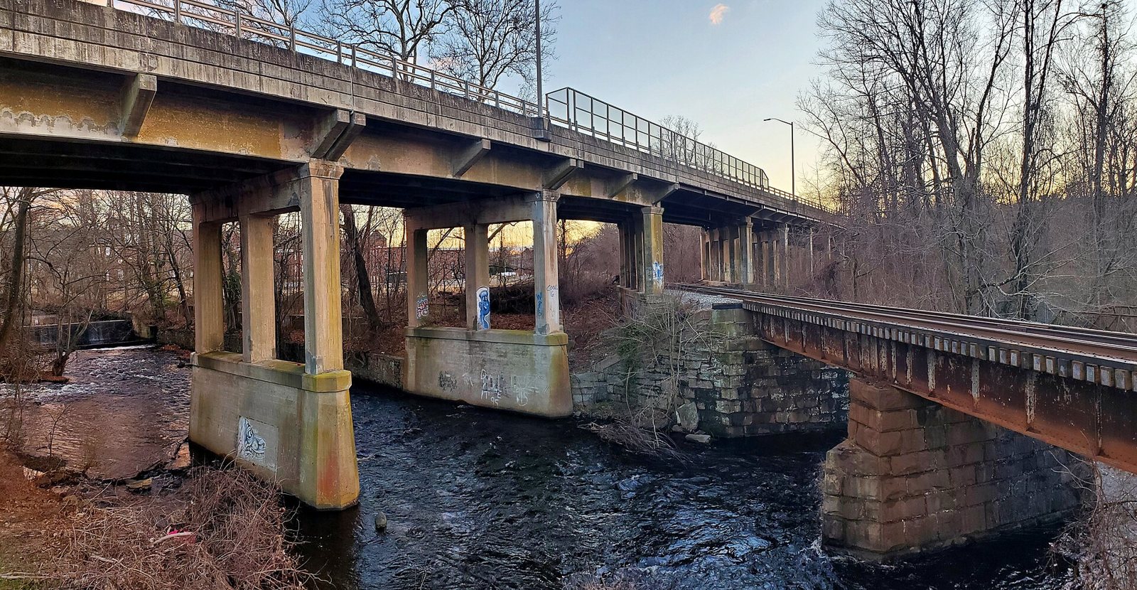 A concrete road bridge and an older railroad bridge on stone piers crossing the Pachaug River in Jewett City, seen from the riverbank