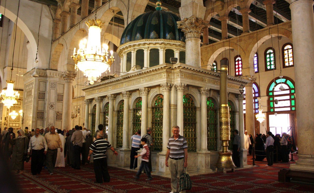 The domed shrine of John the Baptist inside the prayer hall of the Umayyad Mosque, with visitors standing around it under hanging lamps
