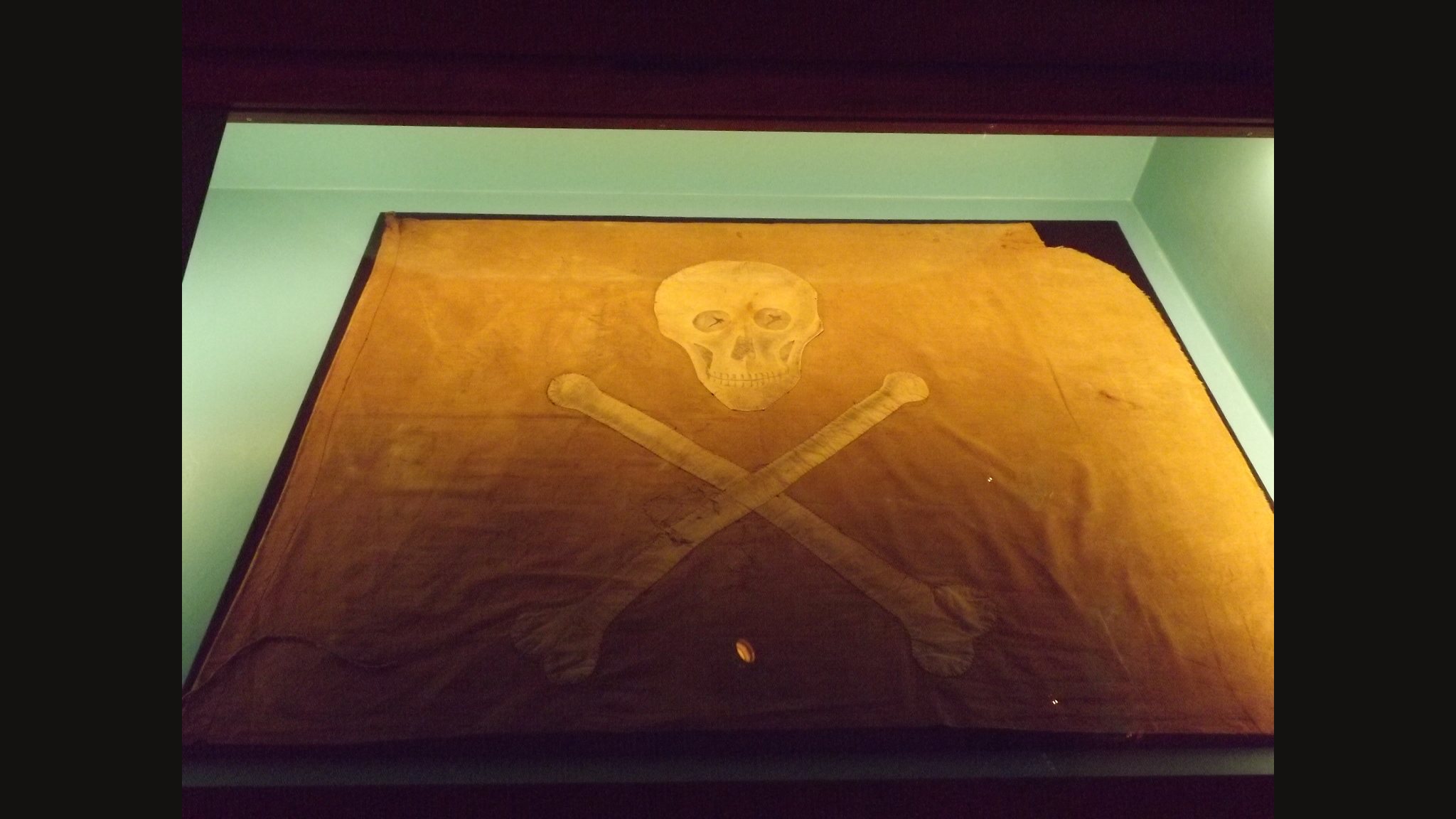 A faded orange-brown flag in a museum case, with a pale cloth skull and two crossed bones stitched to it