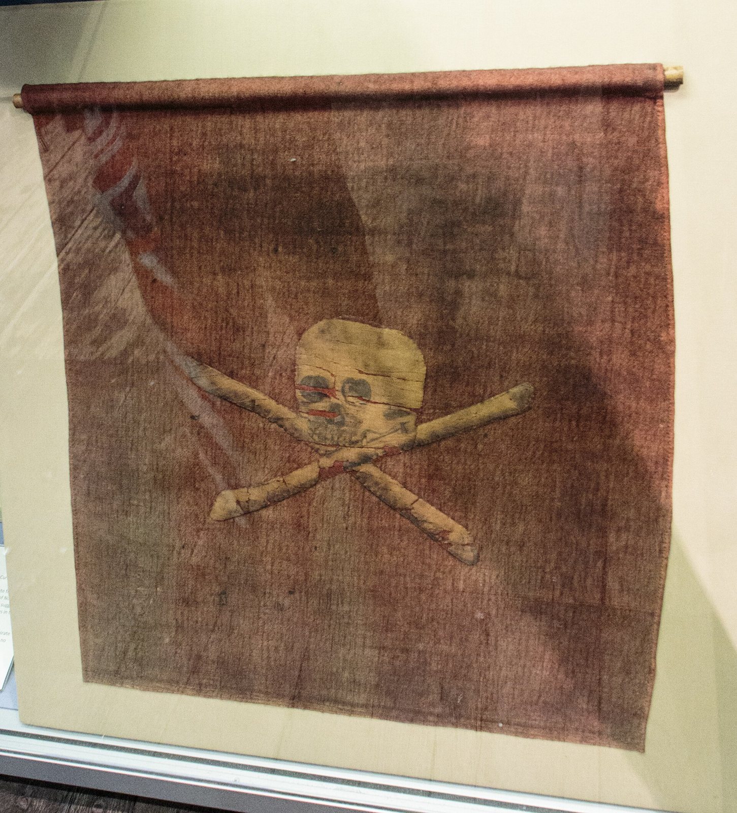 A worn red-brown flag hung from a rod in a display case, with a cream-colored skull and crossed bones at its center