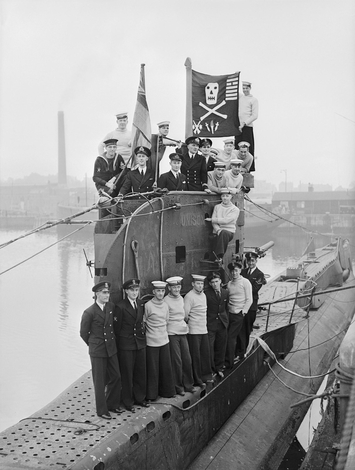 Sailors in jerseys and officers in caps crowd the conning tower and casing of a submarine in harbor, holding up a dark flag with a skull, crossed bones and small symbols