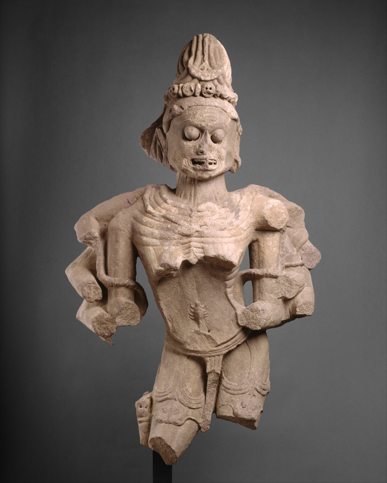 Escultura de arenisca de una diosa demacrada de ojos saltones, dientes al descubierto, costillas talladas en relieve marcado y vientre hundido con un escorpión, una hilera de pequeñas calaveras en su alto tocado y los brazos rotos