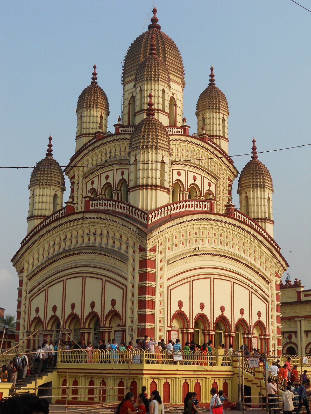 El templo de Kali de Dakshineswar, color crema y rojo, bajo un cielo azul, con sus nueve agujas acanaladas y puntiagudas en niveles sobre una terraza elevada donde se reúnen visitantes a lo lejos