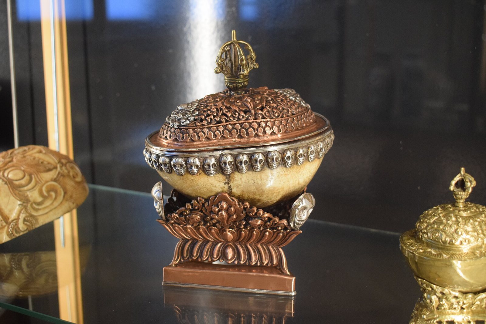 A skull cup with a pierced copper lid, a ring of small silver skulls around its rim and a lotus-shaped copper stand, in a museum case