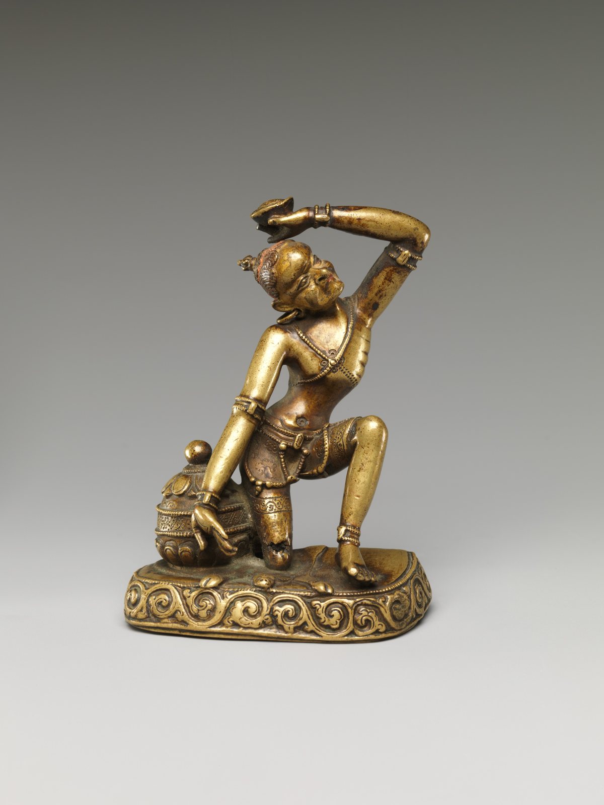 A small gilt brass figure of a kneeling ascetic with one arm raised, holding a skull cup, beside a lidded vessel on a decorated base